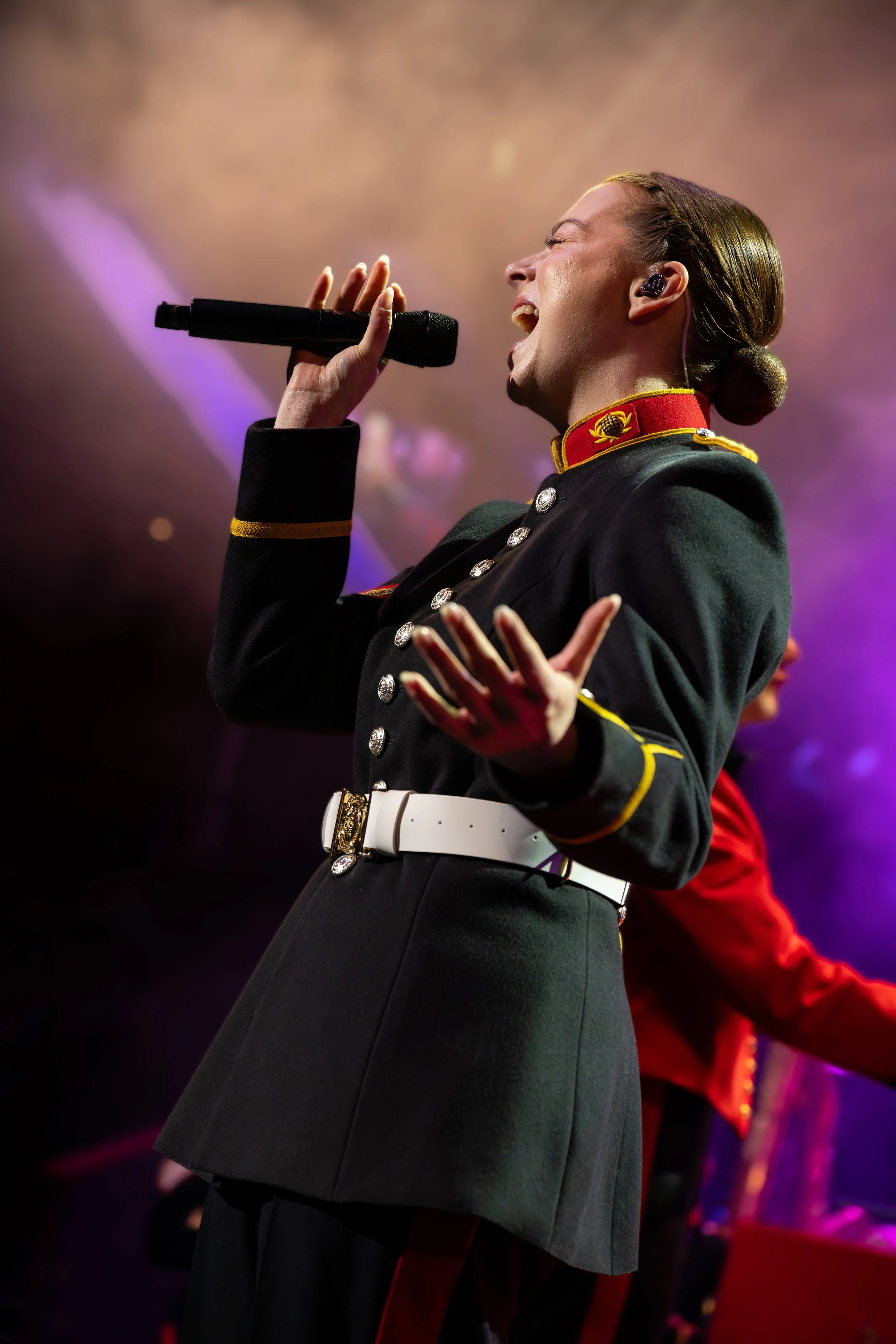 A woman singing into a microphone during a performance, dressed in a military-style uniform with a red collar and gold embellishments, in a concert setting with colorful stage lighting.