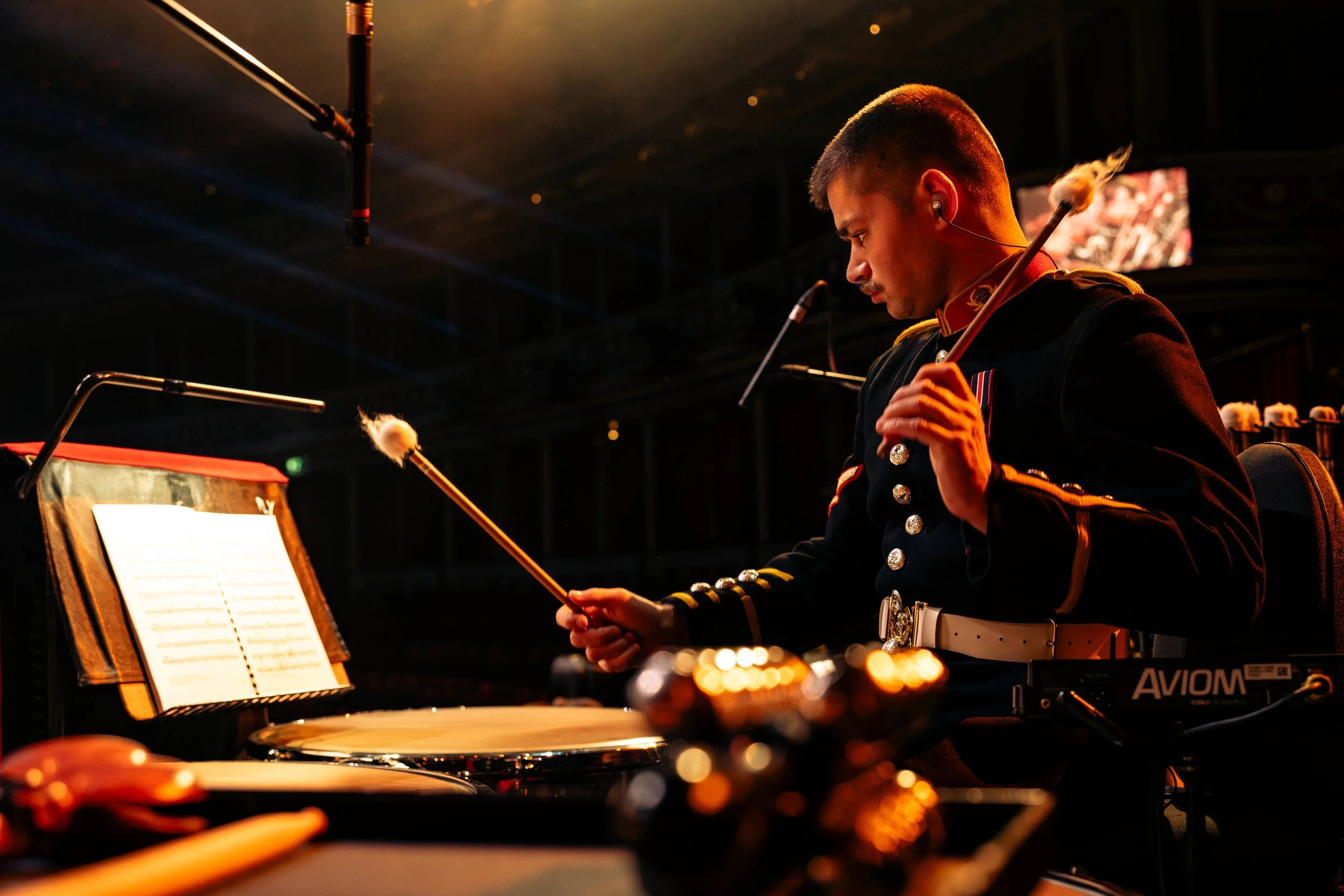 A musician in a military-style uniform playing drums and a xylophone on stage, with sheet music, microphones, and other percussion instruments near him. He is wearing in-ear monitors and is focused on his performance.