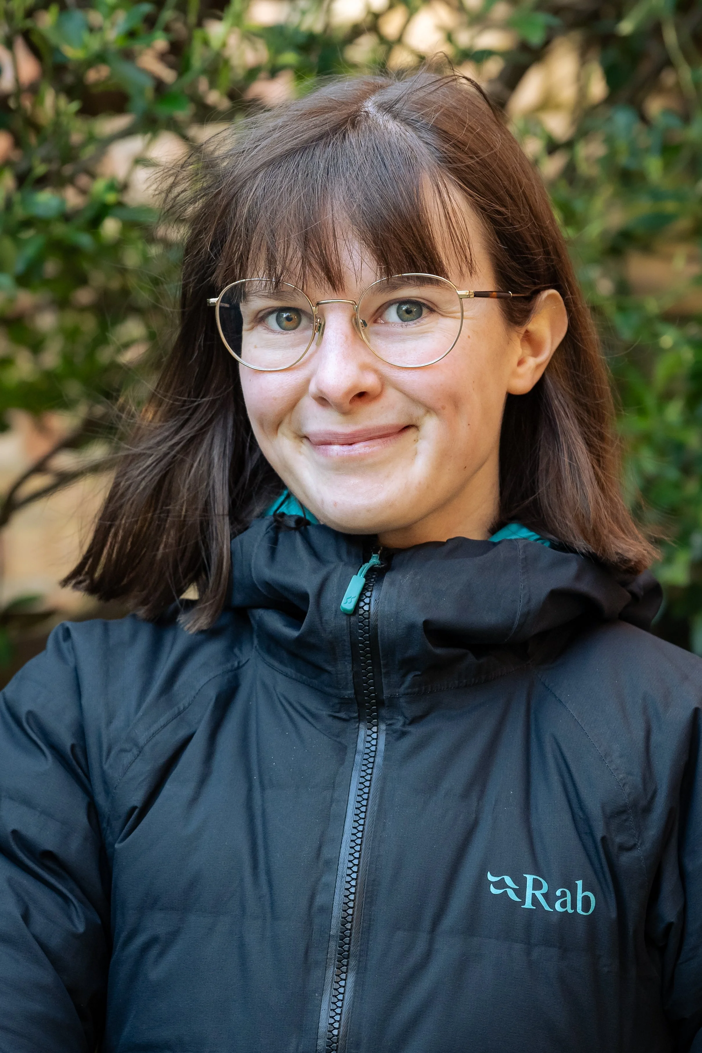 Close-up of a woman with brown hair, glasses, and blue eyes, wearing a black jacket with a logo, standing outdoors with green foliage in the background.