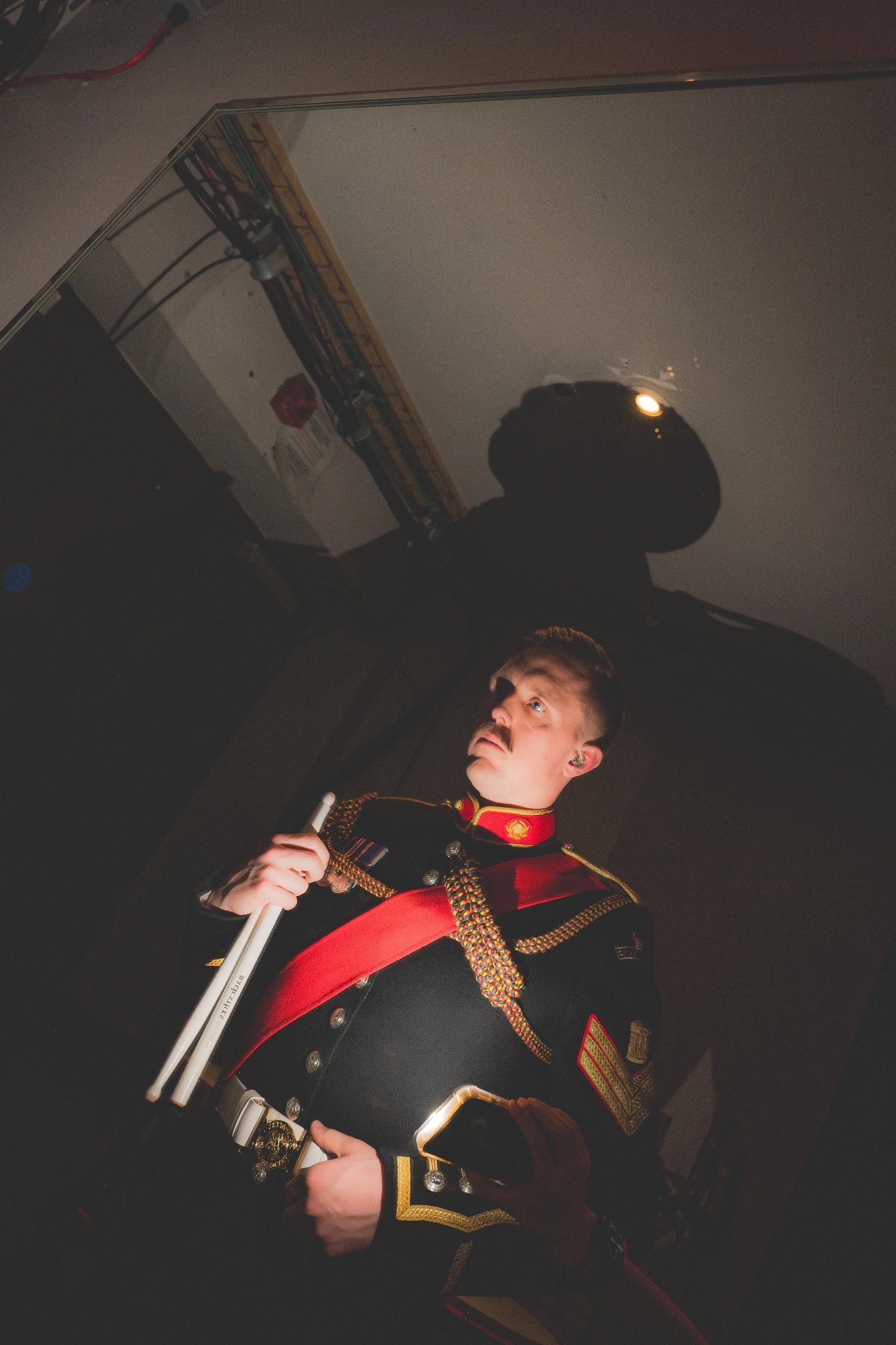A man dressed in a formal military band uniform with a red sash, standing indoors. He has a mustache, and holds a baton in his right hand. The background shows a reflective surface, and some equipment and wires.