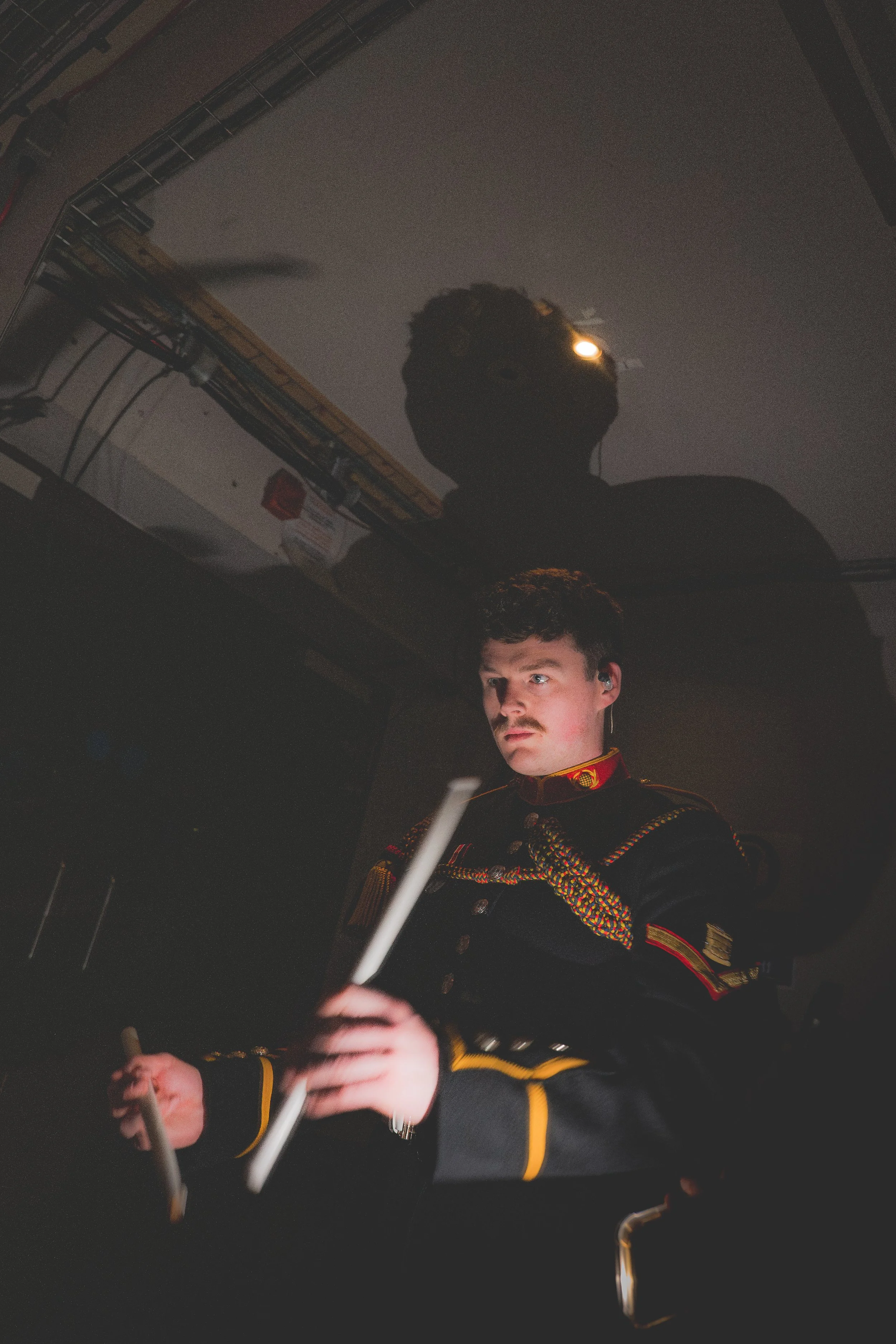 A man in a military uniform with a mustache is looking at a screen, holding a white stick or rod in his hands. His reflection is visible on the shiny surface above him, and there are cables and equipment visible in the background.