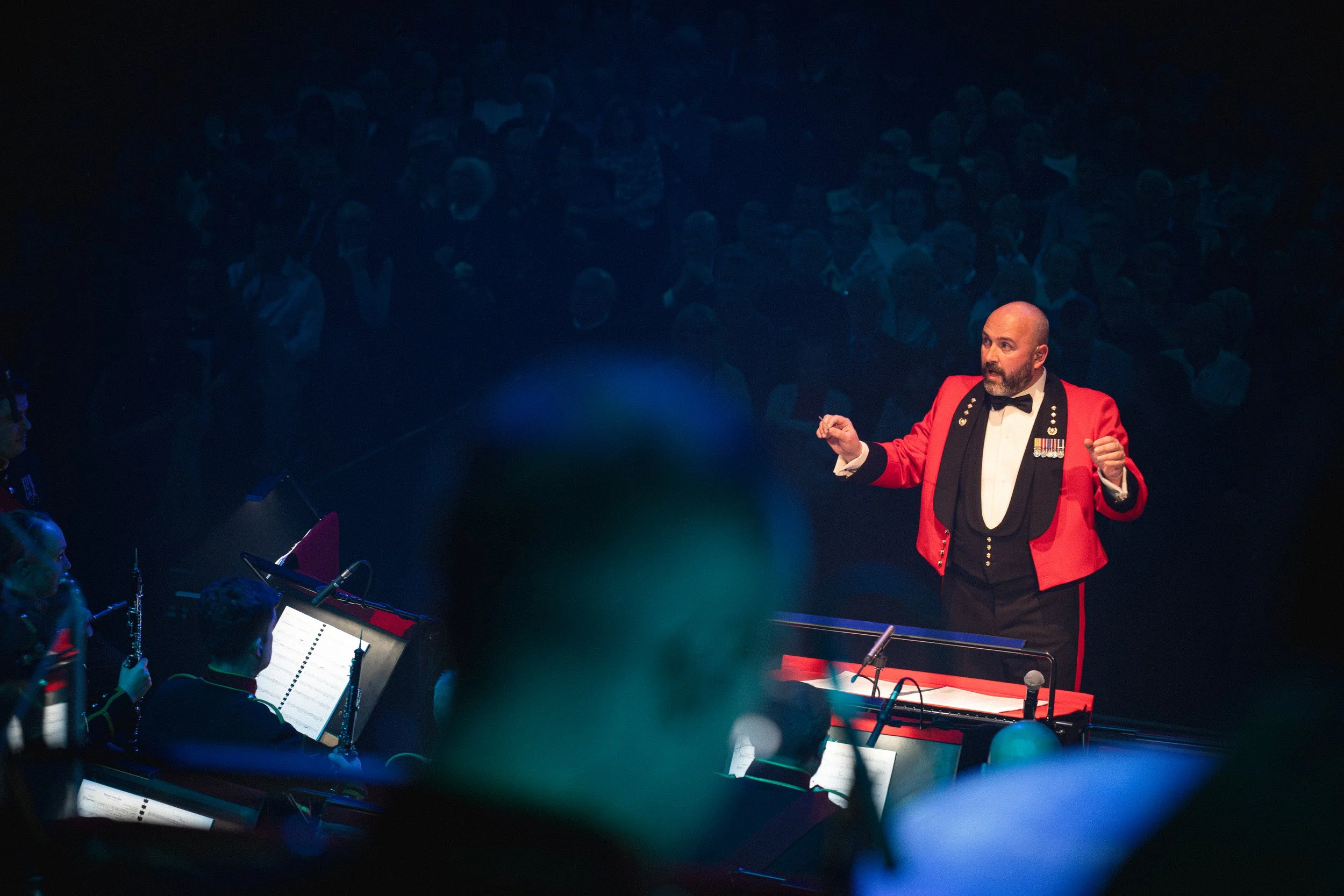 A man in a red tuxedo jacket with medals displays to an orchestra in a dark auditorium.