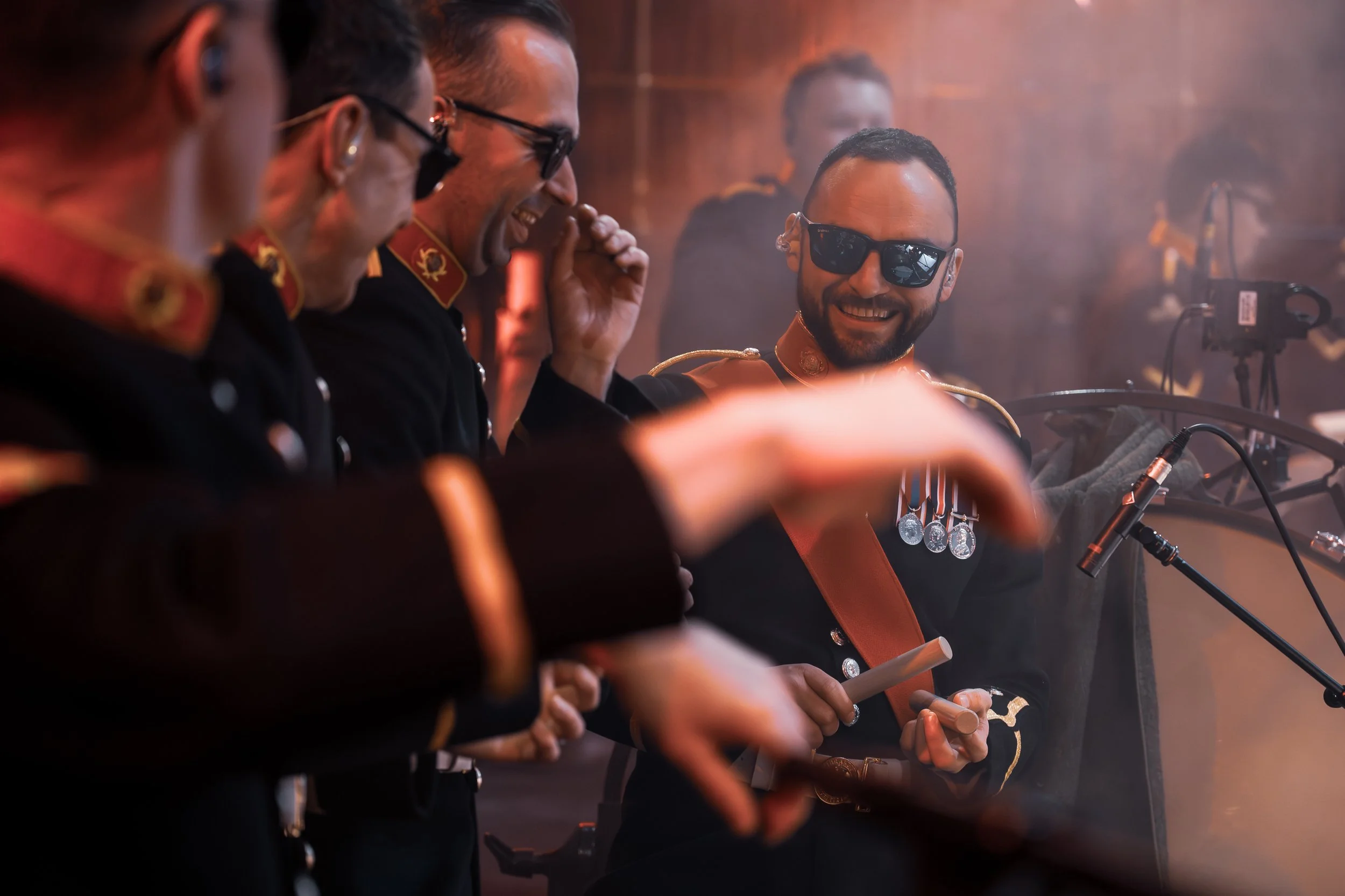 A group of men in military uniforms sitting together, smiling and laughing. They are wearing sunglasses, with some holding and playing musical instruments.