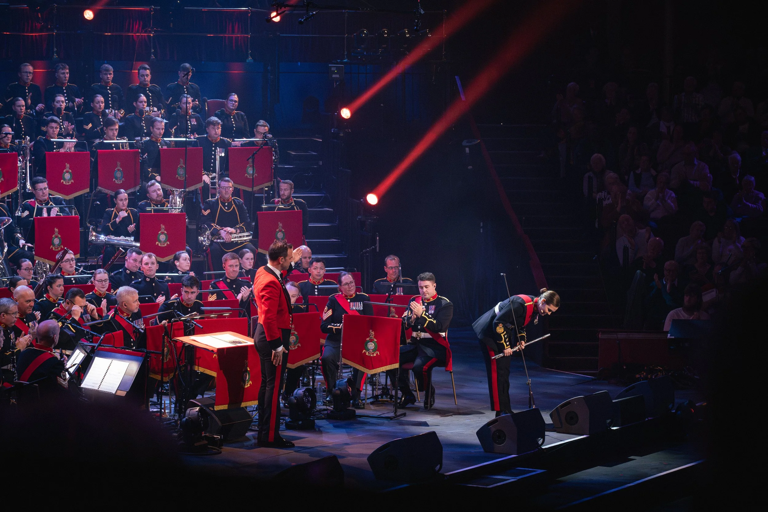 An orchestra performing on stage with musicians in uniform and conductor in a red jacket. Red stage lights and audience in the background.