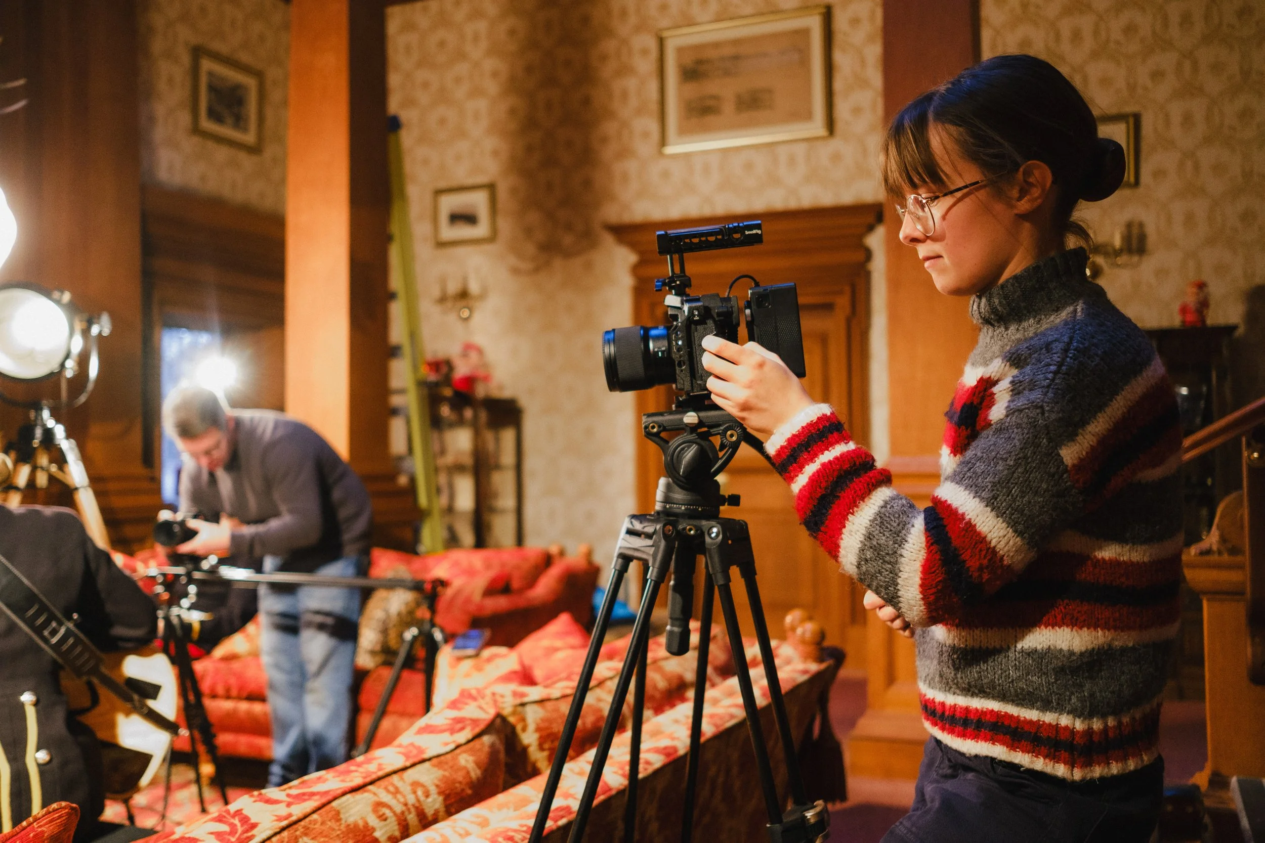 A young girl with glasses adjusting a video camera on a tripod in a warmly lit, vintage-style room with framed pictures on the wall and people preparing equipment.