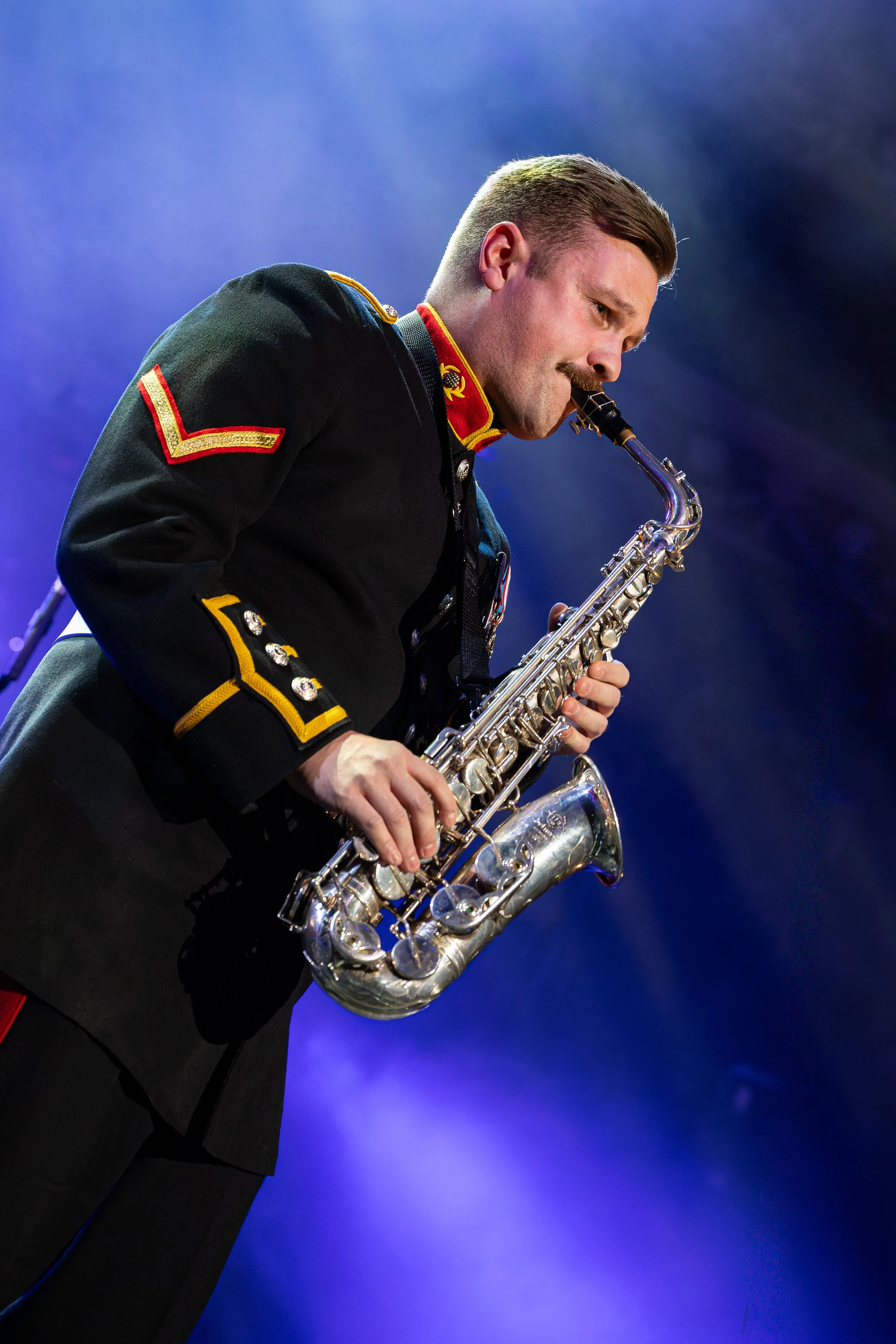 A man in a military uniform playing a silver saxophone on stage with a dark background.