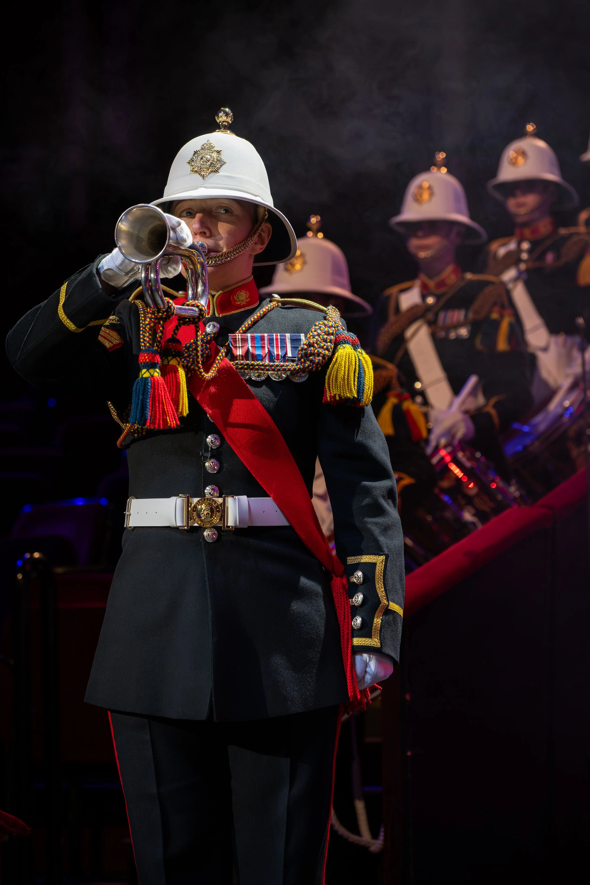 A person dressed as a British soldier playing a trumpet, wearing a traditional uniform with decorative cords, medals, and a white helmet with a badge, in a formation with other similarly dressed individuals in the background.
