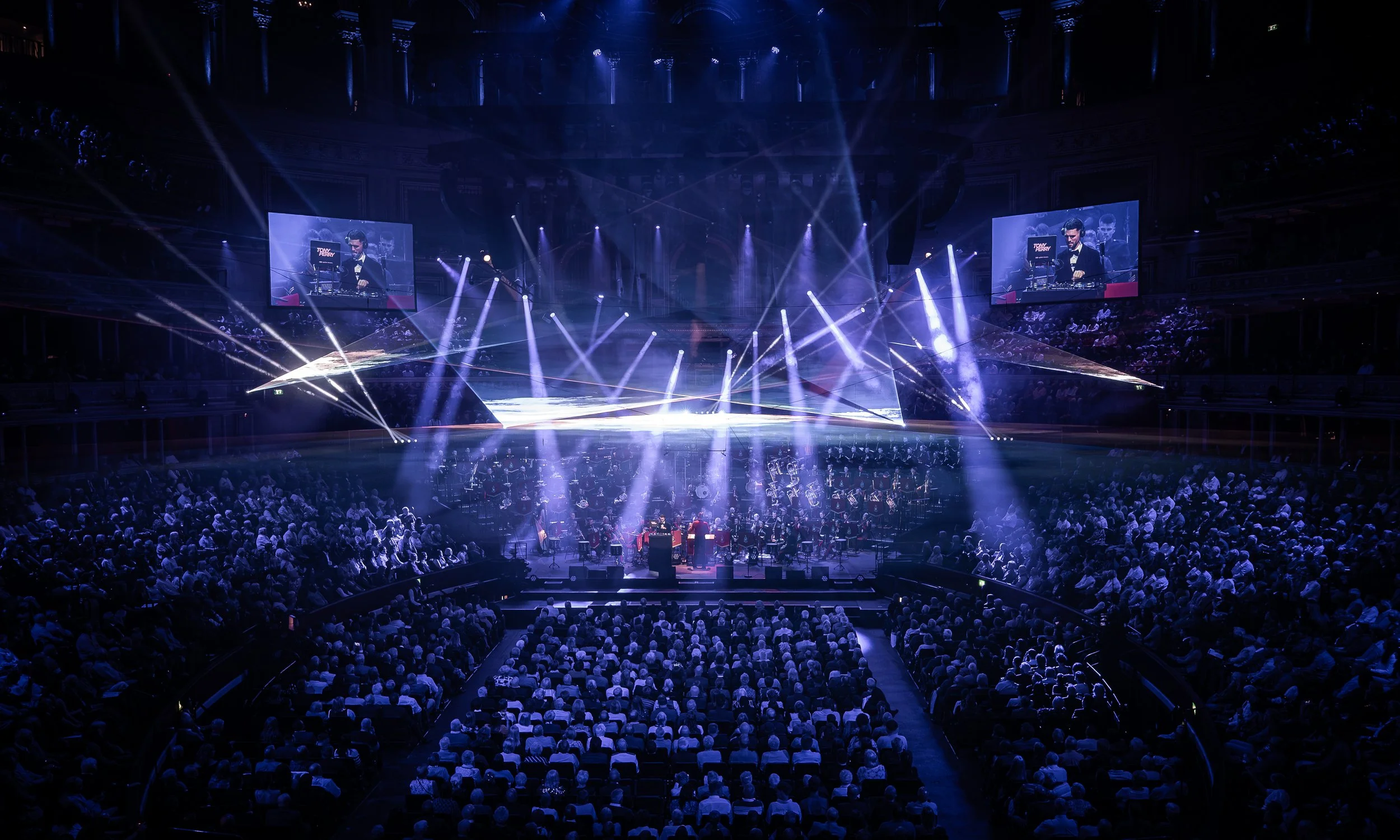 Concert hall with a stage featuring bright lights and large screens displaying a DJ. Audience seated in front, with orchestra sitting below the stage.
