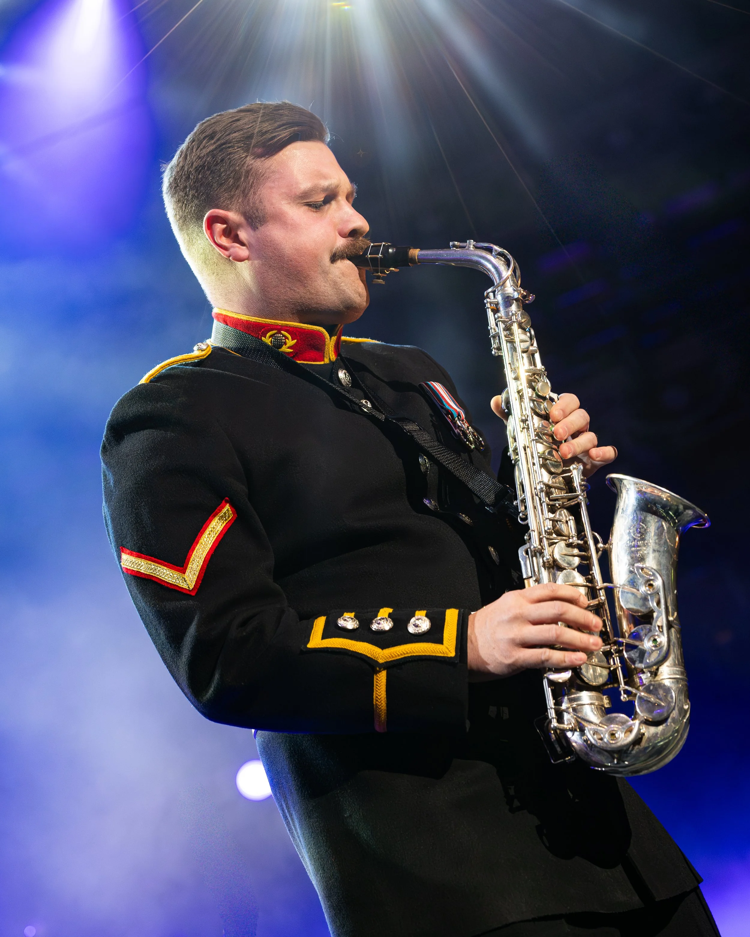 A man in a decorated military uniform playing a saxophone on stage with bright lights shining down.