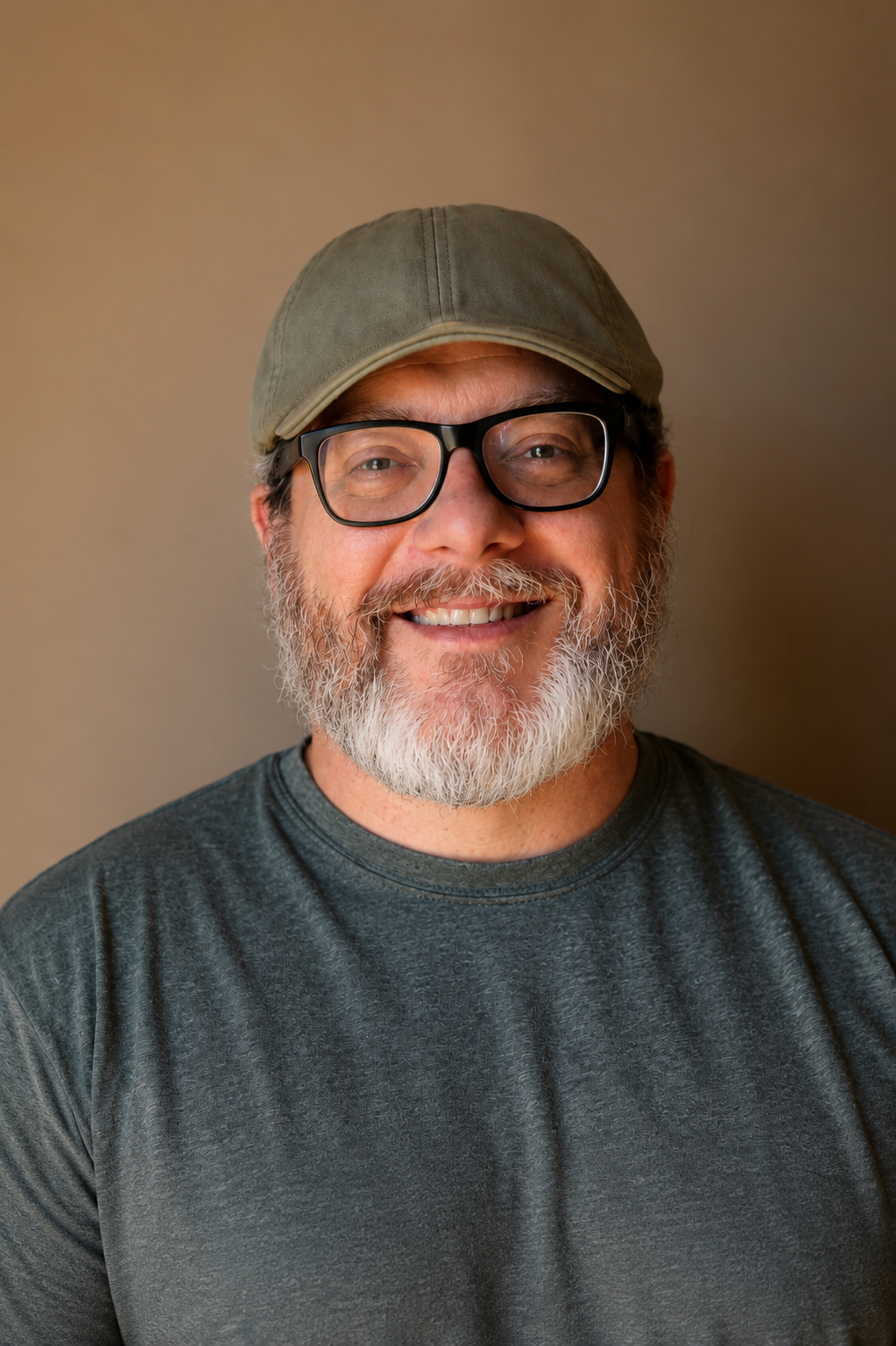 A smiling middle-aged man with a gray beard, glasses, a green cap, and a dark gray shirt against a plain background.