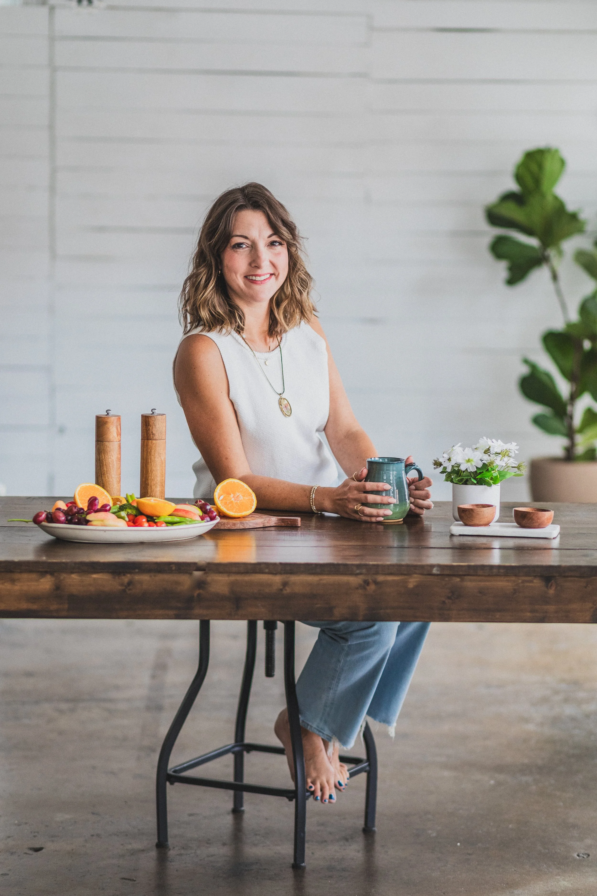 A woman sitting at a wooden table in a white sleeveless top, holding a mug, with a plate of fresh fruit, a potted white flower, and wooden salt and pepper shakers on the table, and a large potted fiddle leaf fig plant in the background.