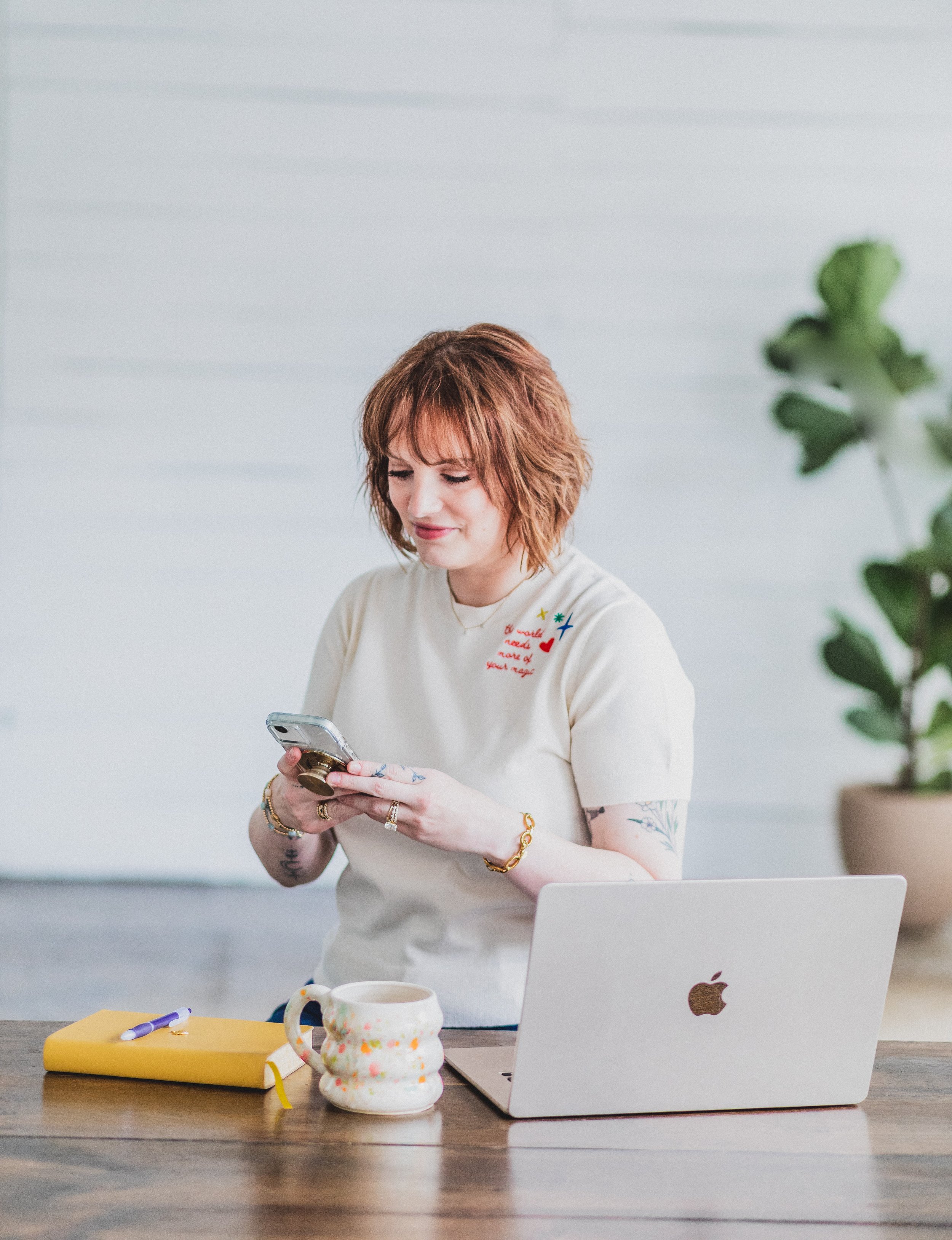 A woman with red hair, wearing a cream-colored T-shirt, stands at a desk looking at her phone with a slight smile. On the desk are a yellow notebook, a purple pen, a ceramic mug, and a silver laptop with an Apple logo. In the background, there's a large green plant and a light-colored wall.