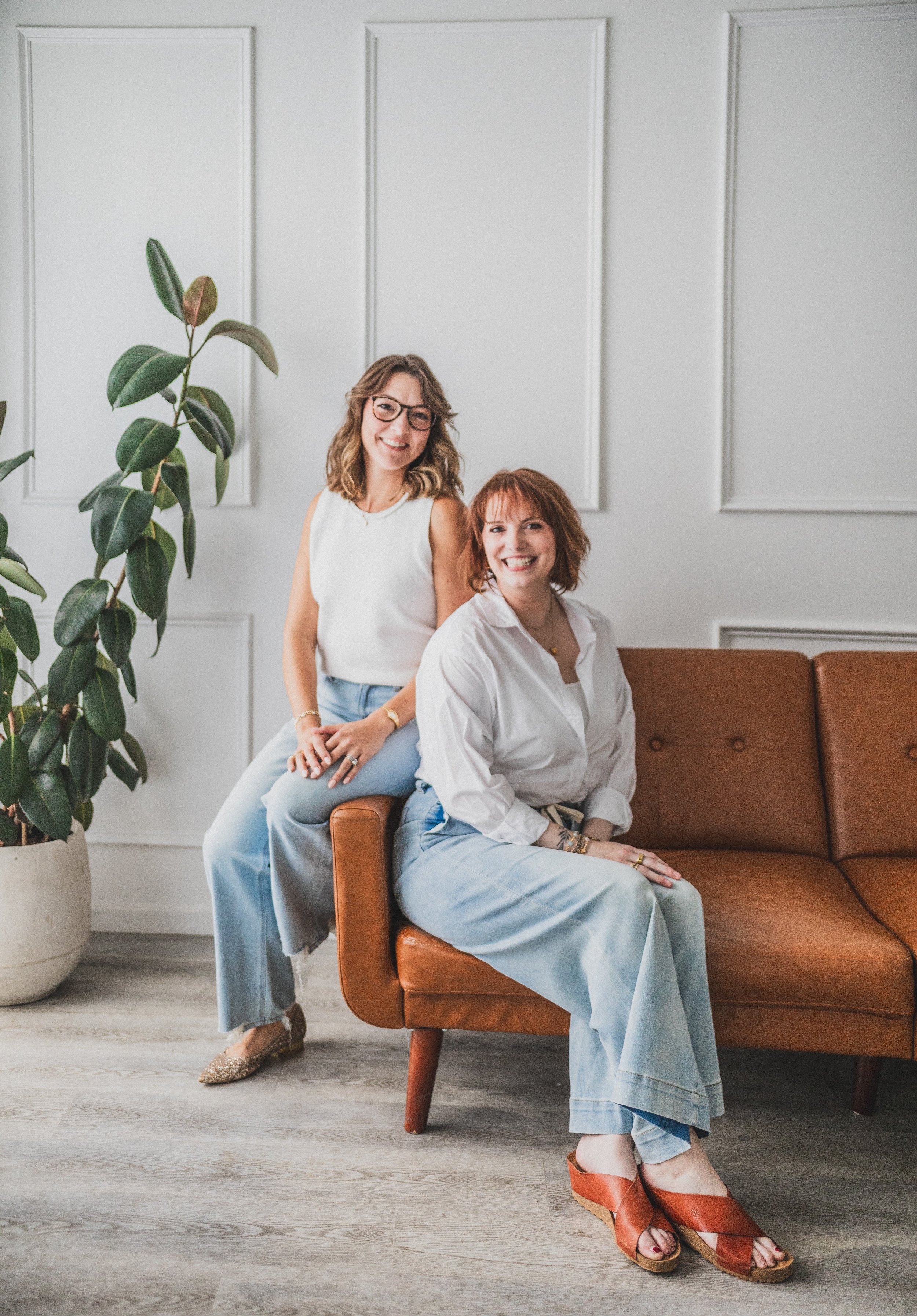 Two women sitting and standing near a brown couch in a modern, well-lit living room with a large plant and decorative wall panels.