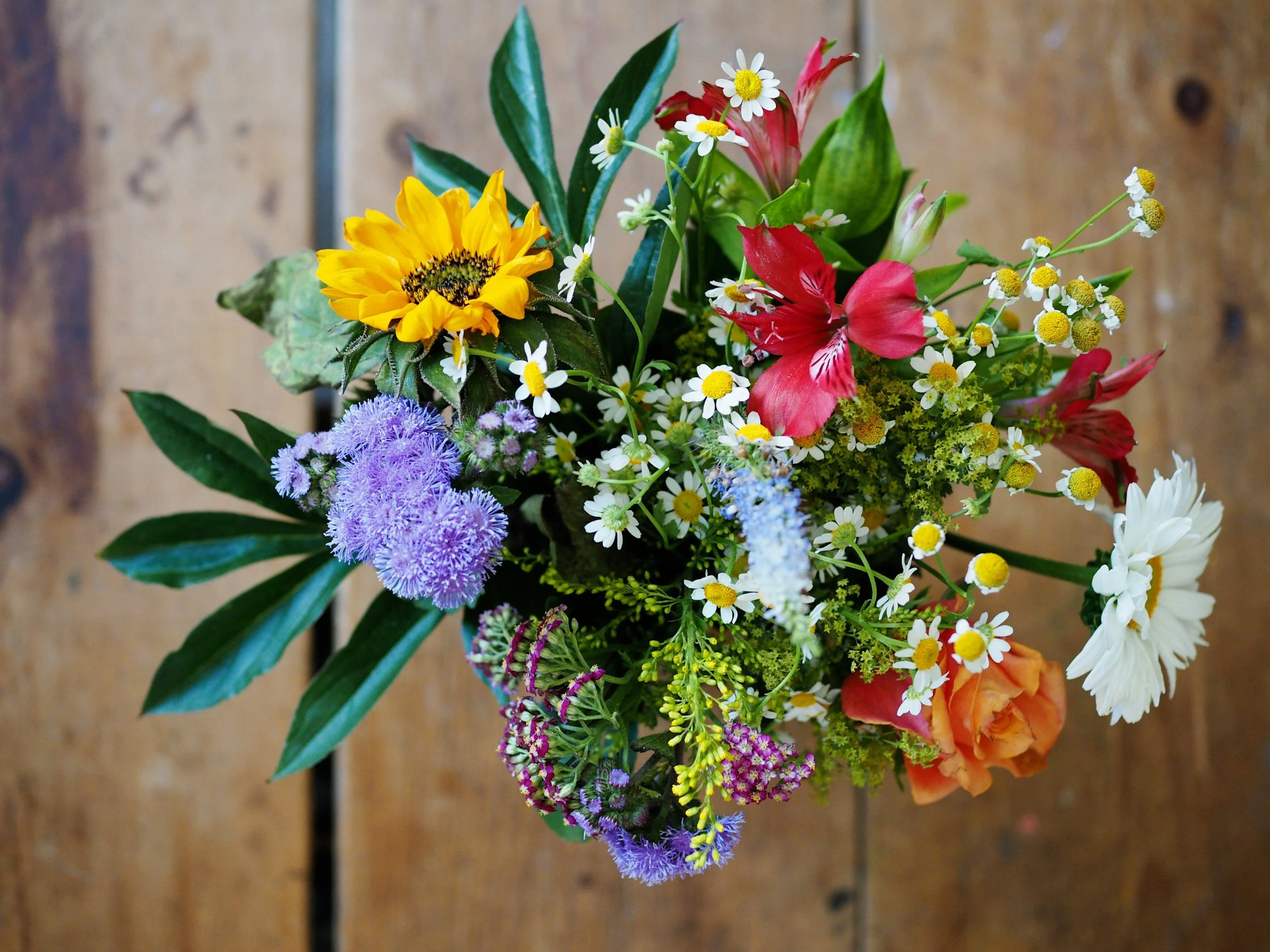 A bouquet of mixed flowers including a sunflower, daisies, pink lilies, purple asters, and orange blooms, with green leaves against a wooden background.