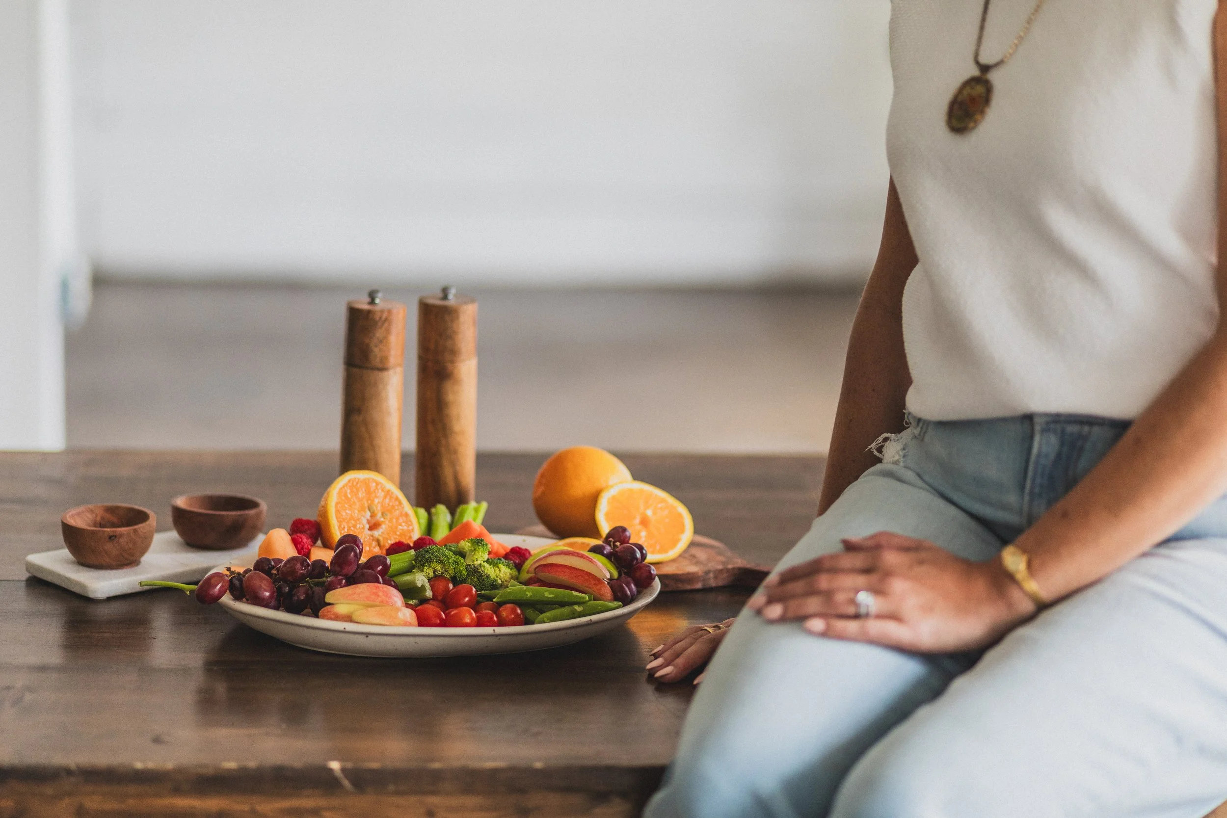 A woman sitting next to a wooden table with a plate of fresh fruits and vegetables, including grapes, strawberries, cherry tomatoes, broccoli, and sliced oranges, with pepper mills and small bowls in the background.