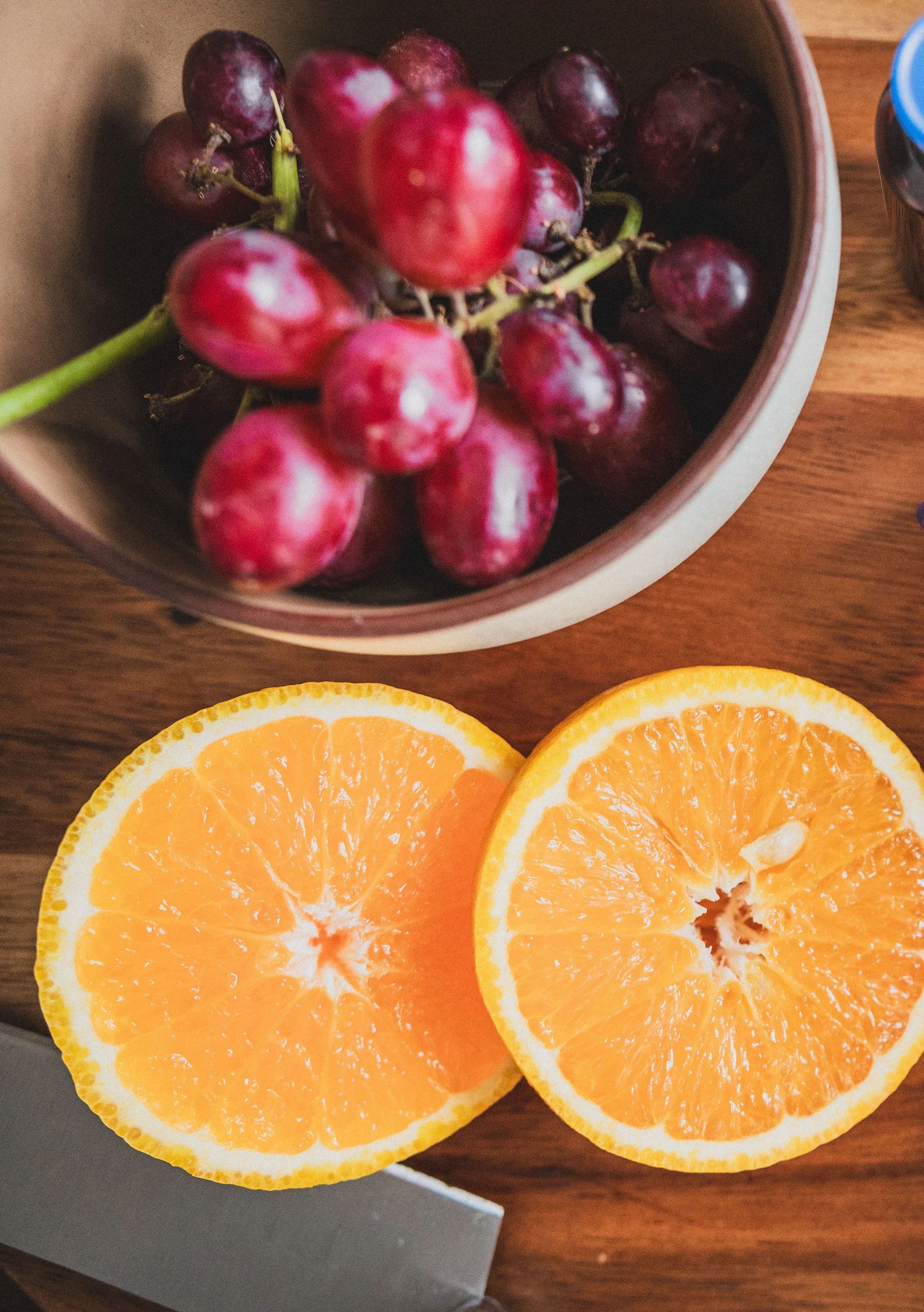 A bowl of red grapes and two halves of an orange on a wooden surface.
