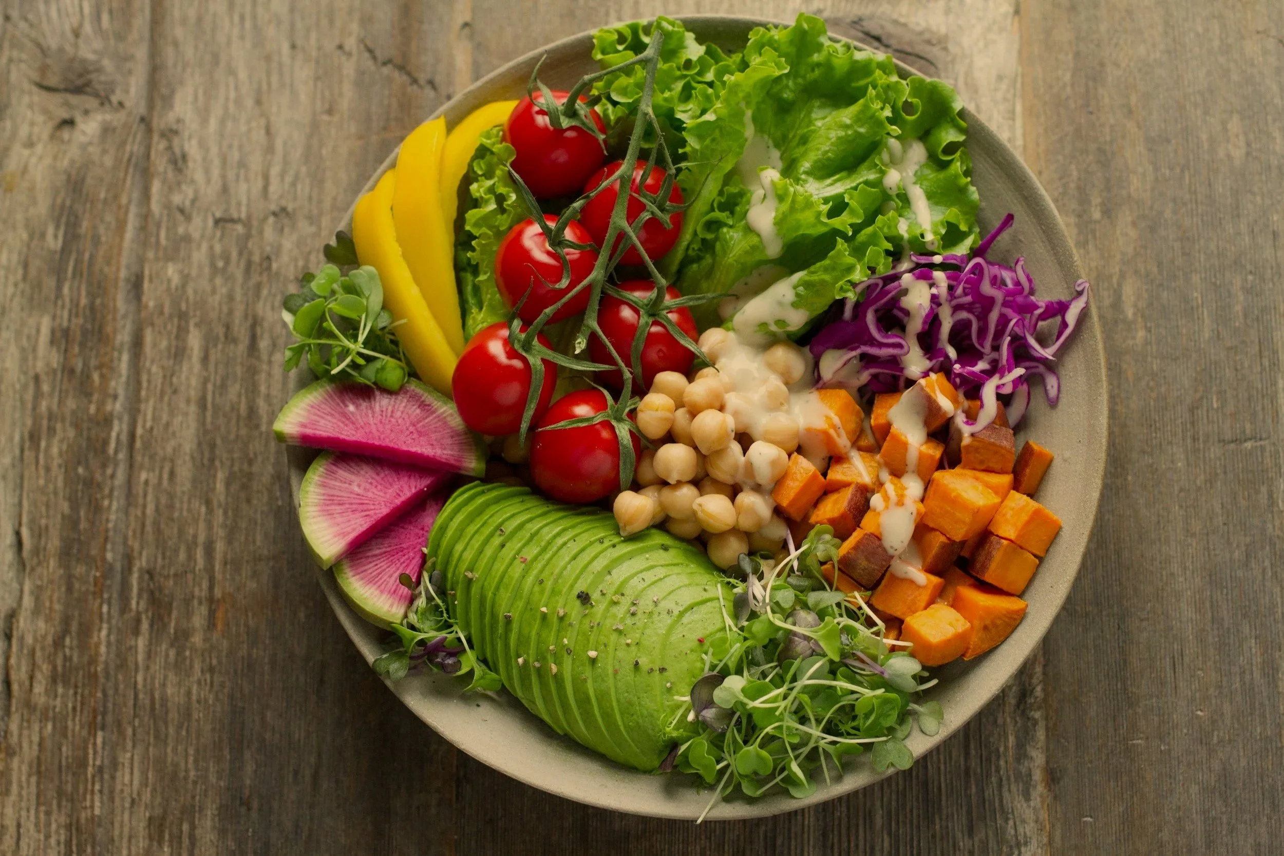 Assorted fresh vegetables and greens in a bowl on a wooden surface, including cherry tomatoes, sliced avocado, watermelon radish, chopped sweet potatoes, shredded purple cabbage, chickpeas, greens, and sliced yellow zucchini.