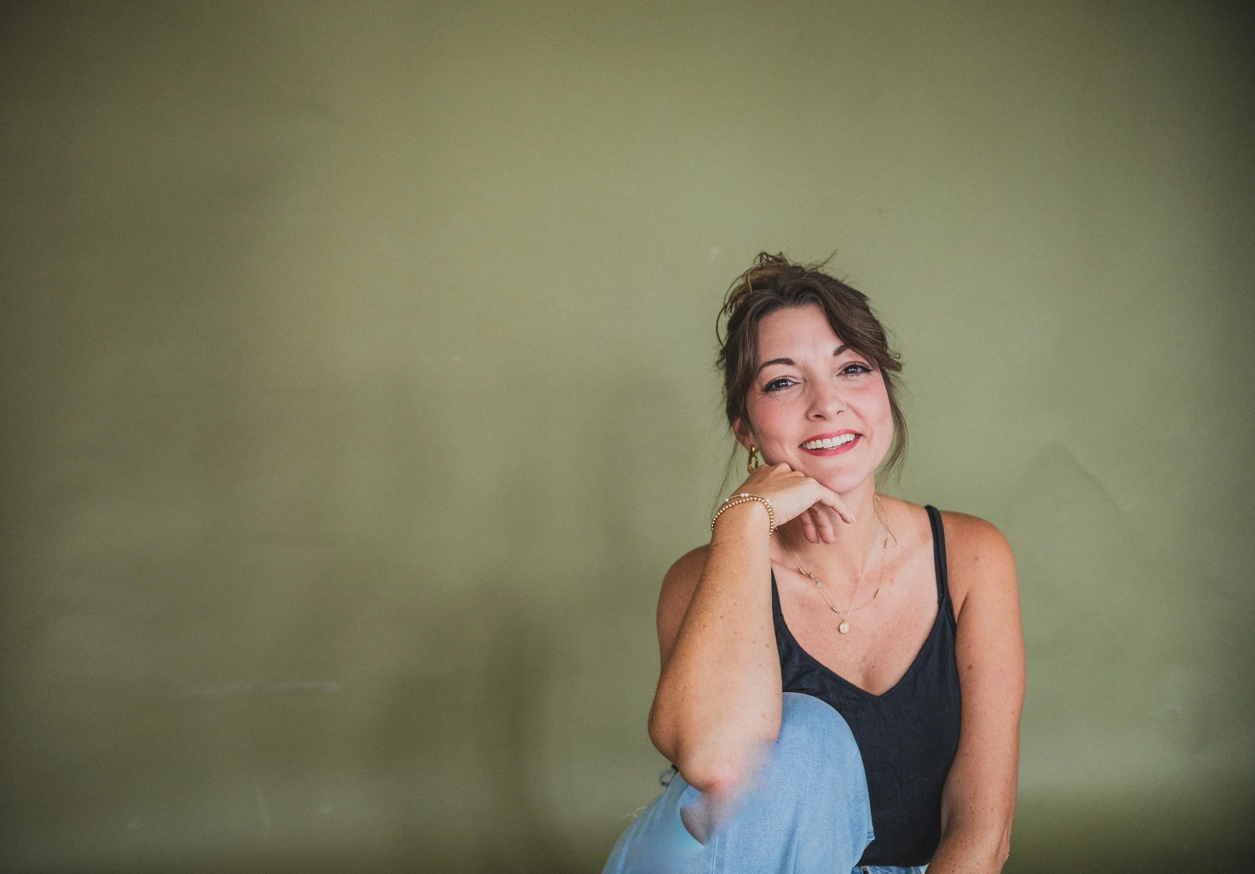 A woman with brown hair and freckles sits against a plain green wall, smiling at the camera with her chin resting on her hand. She wears a black sleeveless top, gold earrings, and layered gold necklaces.