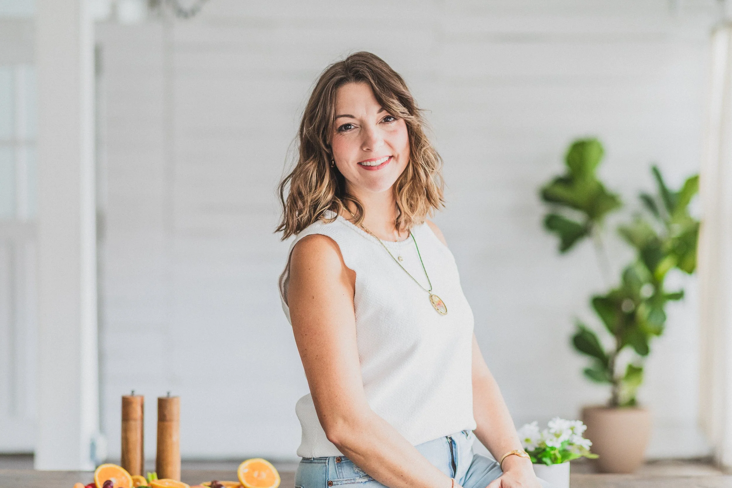 A woman with wavy brown hair, wearing a white sleeveless top and light blue jeans, smiling at the camera in a bright, modern kitchen with potted plants and fruit on the countertop.