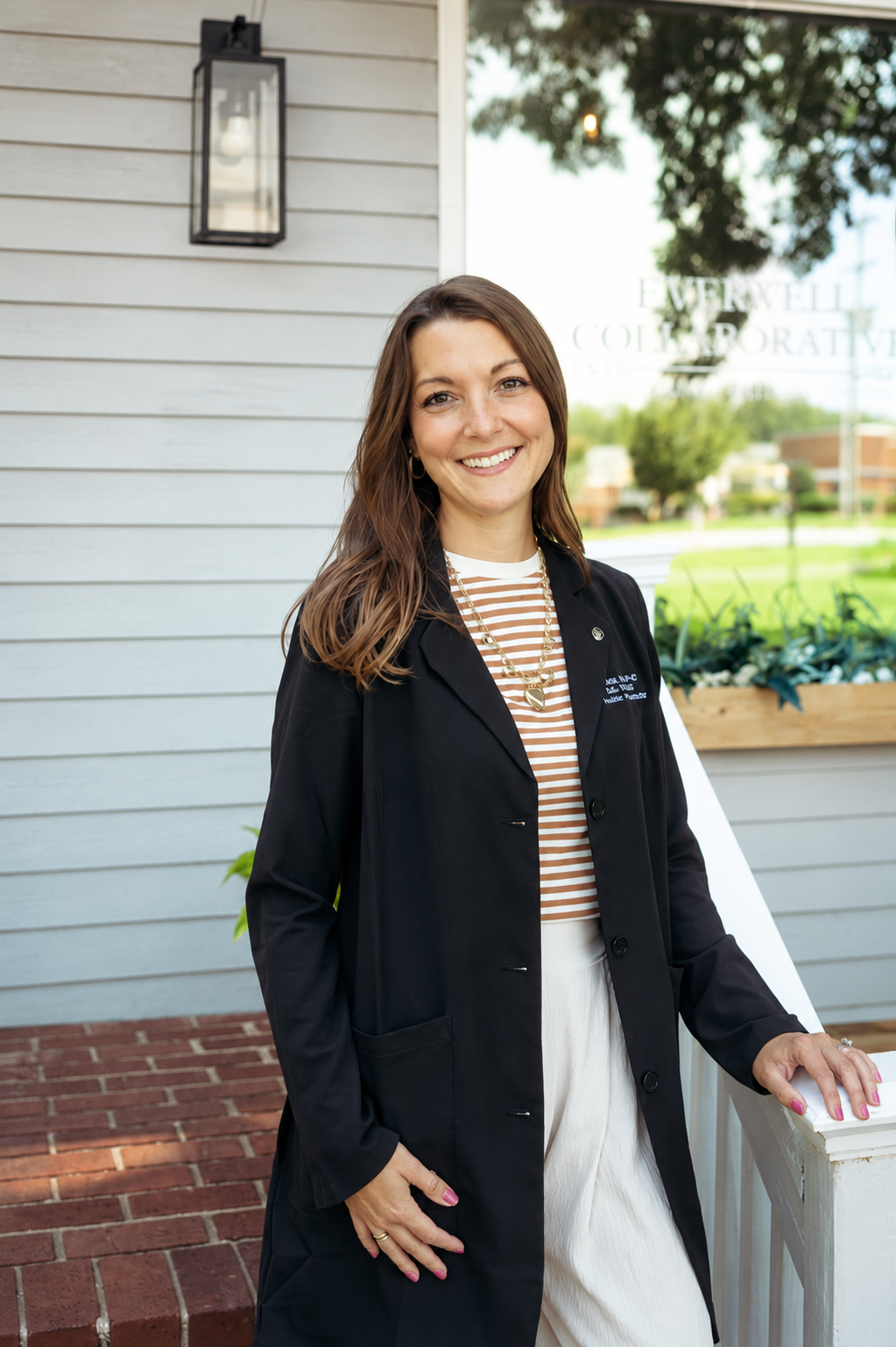 A smiling woman with brown hair standing outside a building, dressed in a black blazer over a striped shirt and white skirt, with a gold necklace, in front of a window and house siding.
