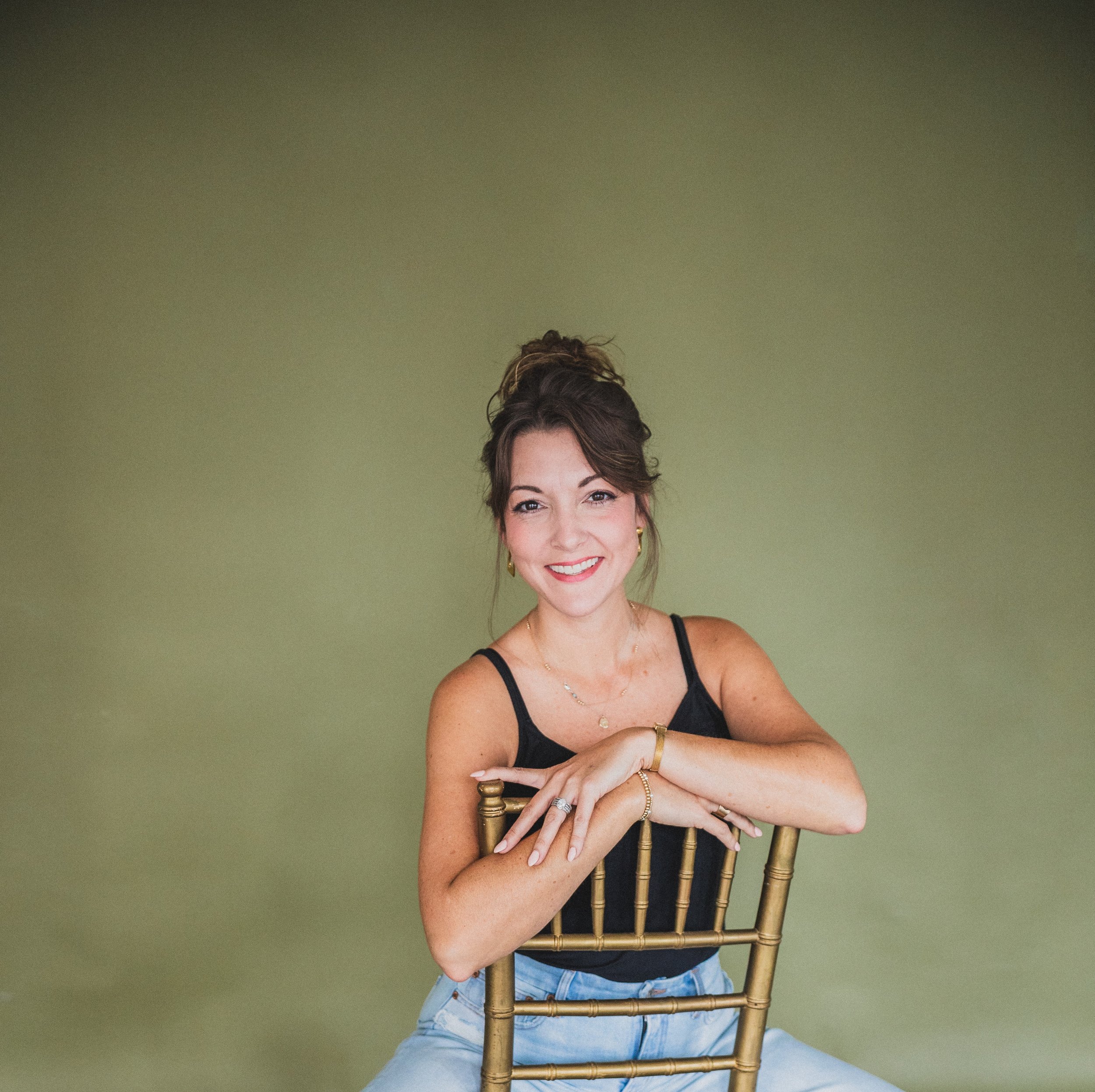 A woman with dark hair styled in an updo, wearing a black tank top and gold jewelry, sits on a gold chair against a light green background, smiling at the camera.