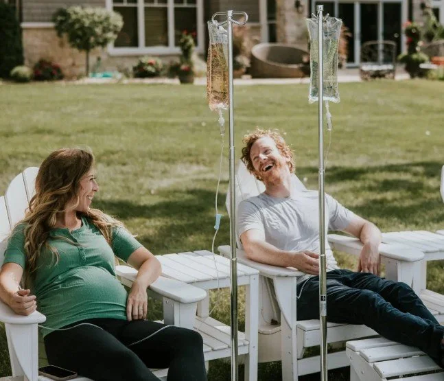 A woman and a man sitting on outdoor white chairs, laughing, with IV poles and bags of fluids attached to them, outdoors in a garden.