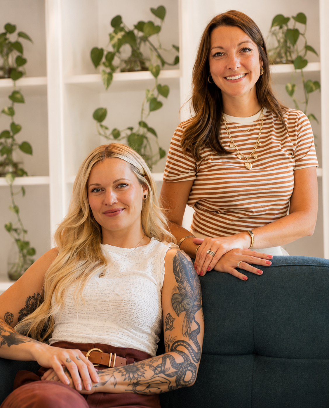 Two women in a living room with green plants on white shelves. One woman is seated on a couch with tattoos on her arms, wearing a white sleeveless top. The other woman is standing behind her, wearing a striped t-shirt, smiling with her hand resting on the back of the couch.