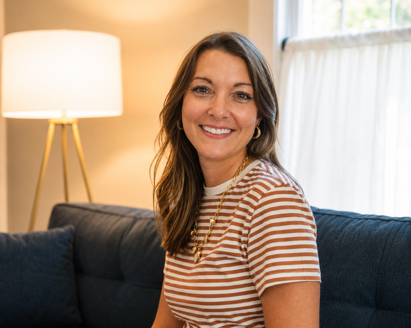 A woman with long brown hair, dressed in a striped shirt and gold jewelry, smiling while sitting on a dark blue couch in a warmly lit living room near a window.