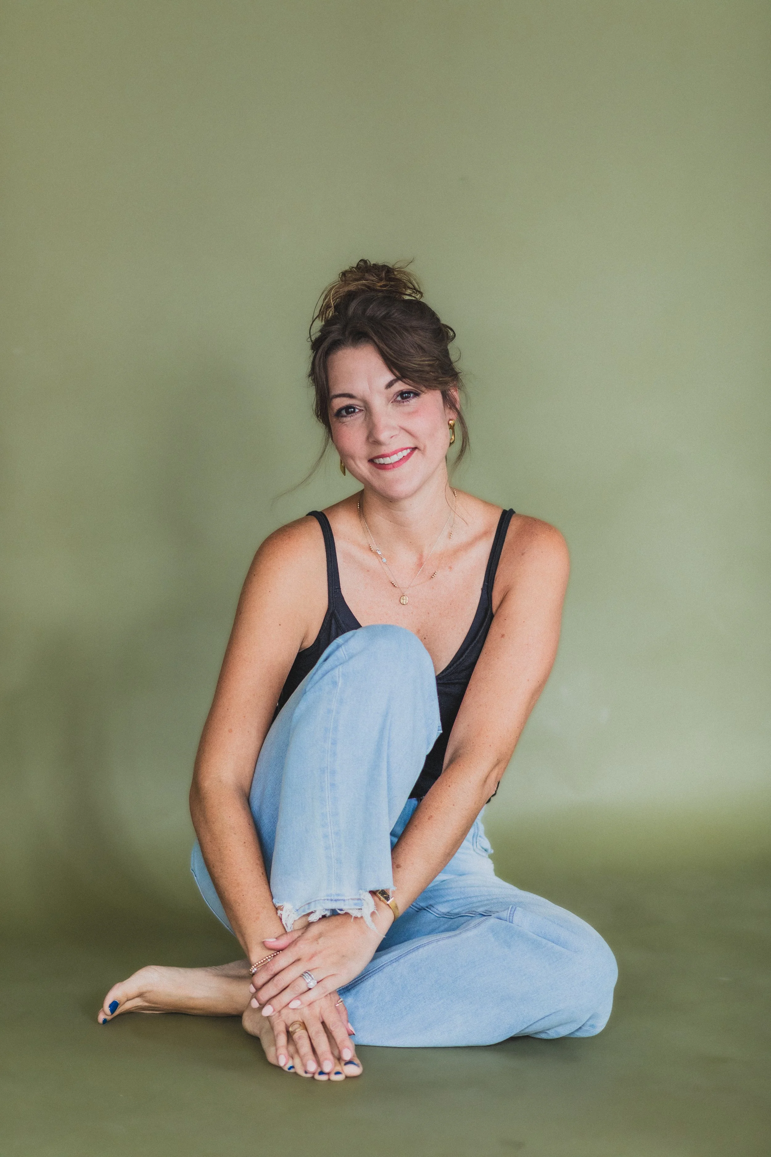 A woman sitting on the floor against a plain green background, smiling, with her hair in a messy bun, wearing a black tank top and light blue jeans.