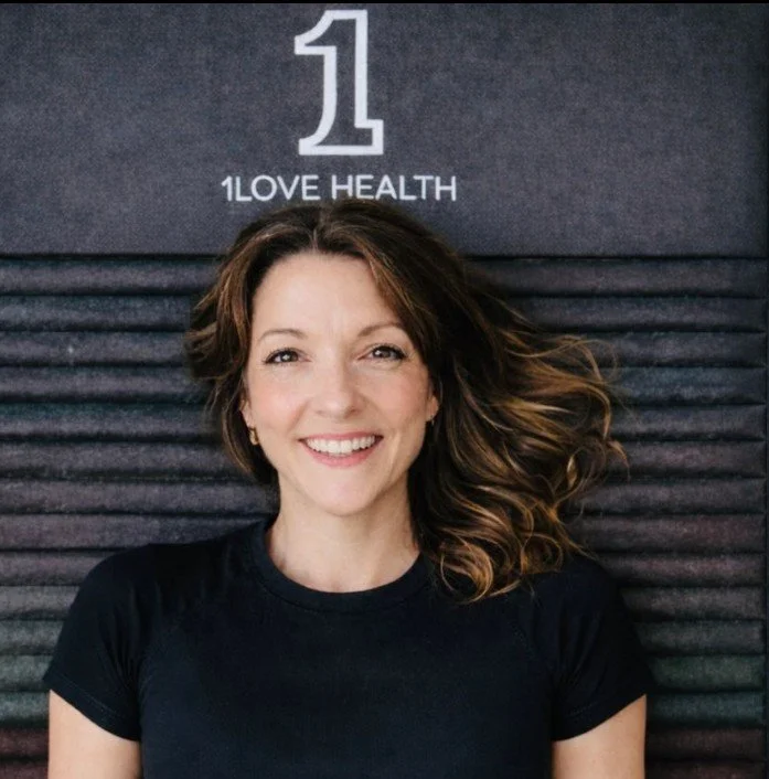 A woman with wavy brown hair and a black shirt smiling in front of a dark background with the text '1 LOVE HEALTH' at the top.
