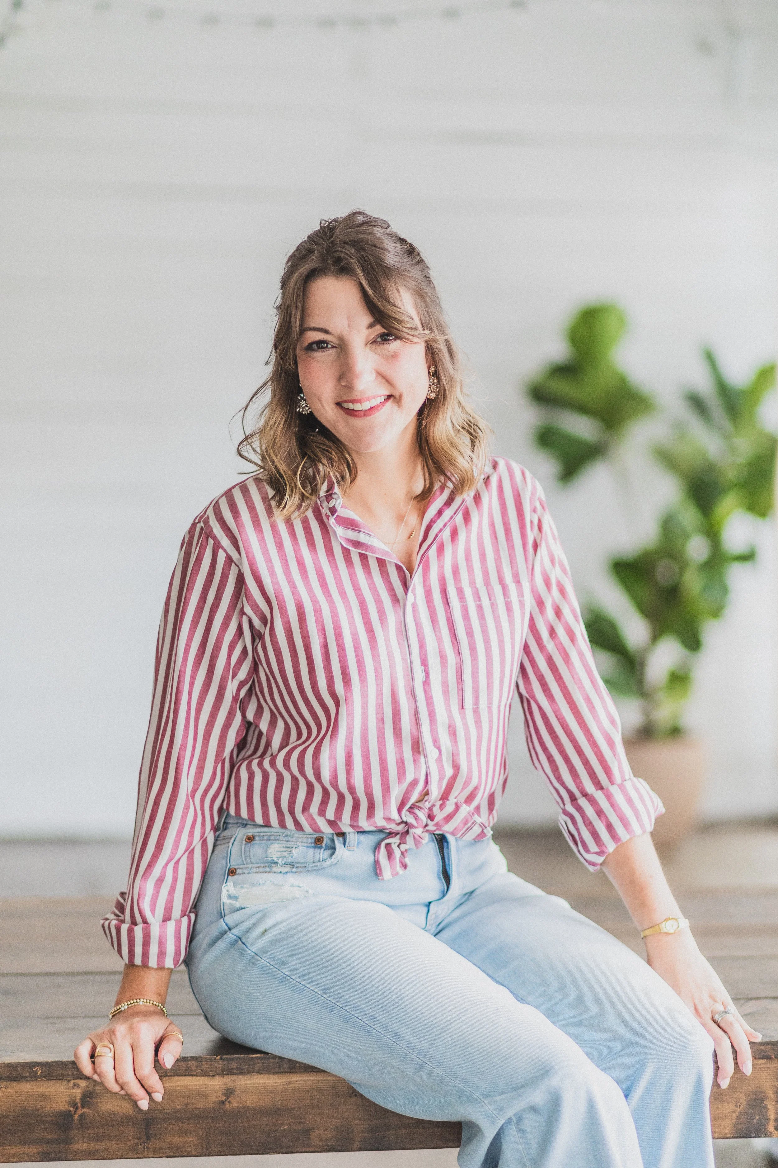 A woman with shoulder-length wavy brown hair, wearing a pink and white striped shirt tied at the waist, and light blue jeans, sitting on a wooden surface indoors with a green potted plant in the background, smiling at the camera.