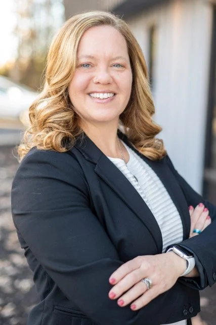 A woman with blonde, curly hair smiling with arms crossed, wearing a dark blazer and a striped shirt, standing outdoors.