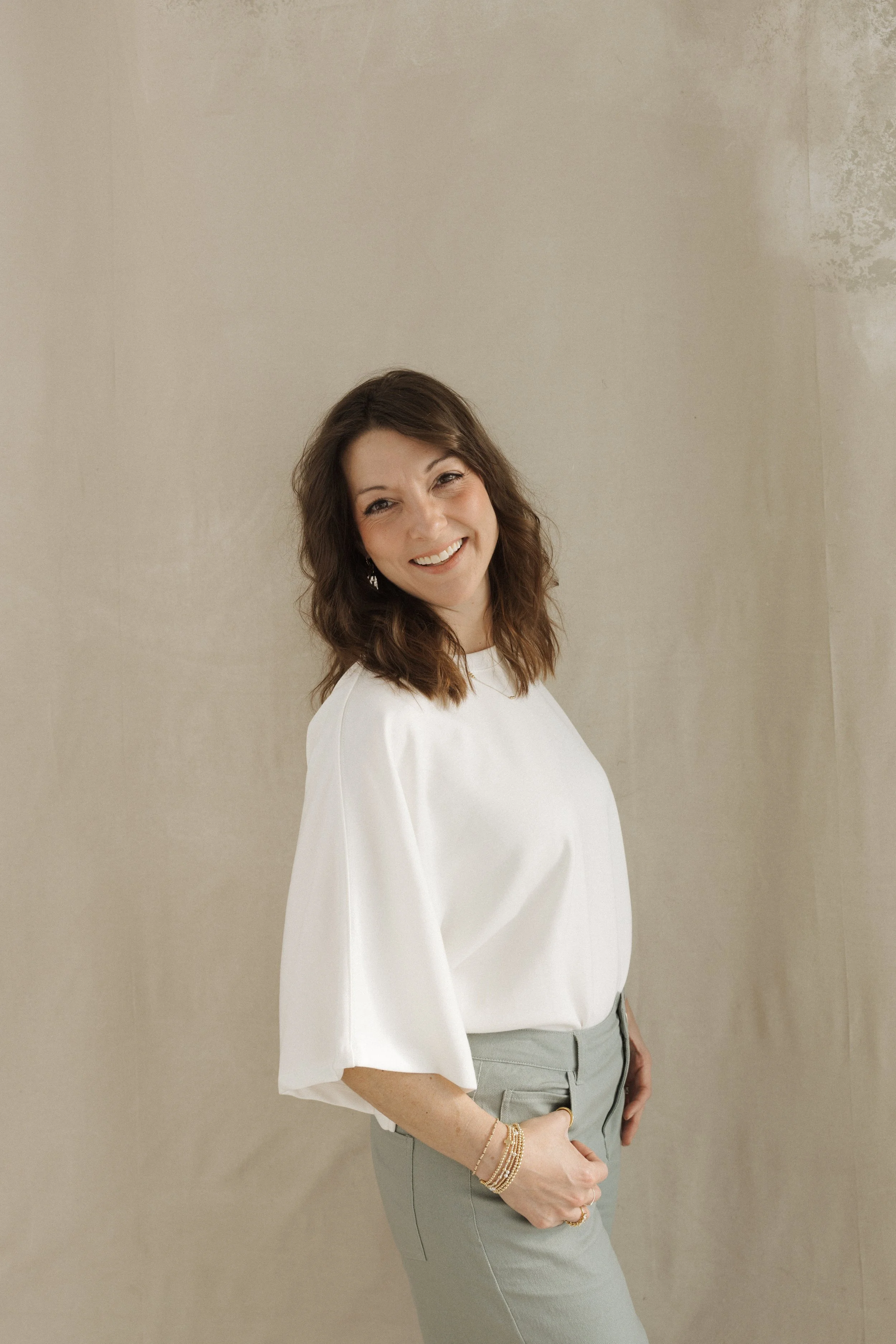 Woman with shoulder-length brown hair smiling, wearing a white top with wide sleeves and gray pants, standing against a plain beige wall.