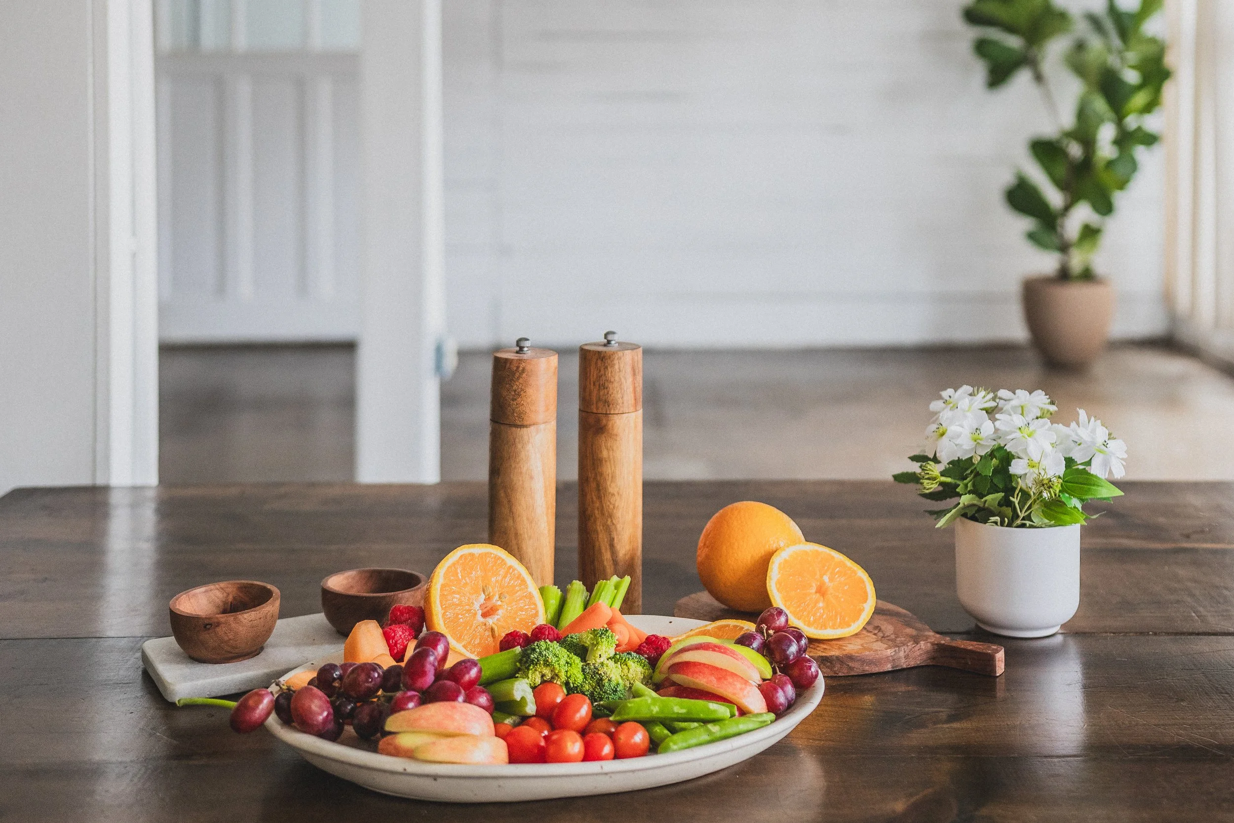 A wooden table with a white bowl of assorted fruits including grapes, cherry tomatoes, apple slices, broccoli, carrots, and orange halves. There are two wooden salt and pepper shakers, a small wooden bowl, and a white potted plant with white flowers in the background.