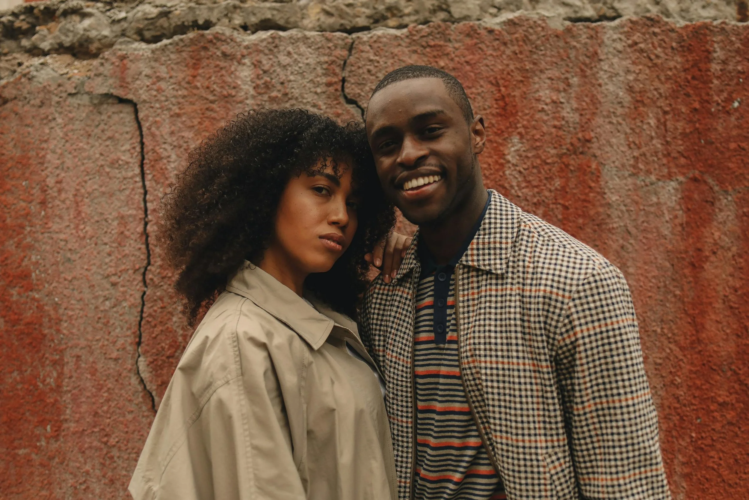 A woman and a man standing close together in front of a red textured wall, with the woman's arm resting on the man's shoulder. The woman has curly hair and is wearing a beige jacket. The man has short hair, dark skin, and is wearing a checkered shirt over a striped polo shirt. They are both smiling and looking at the camera.