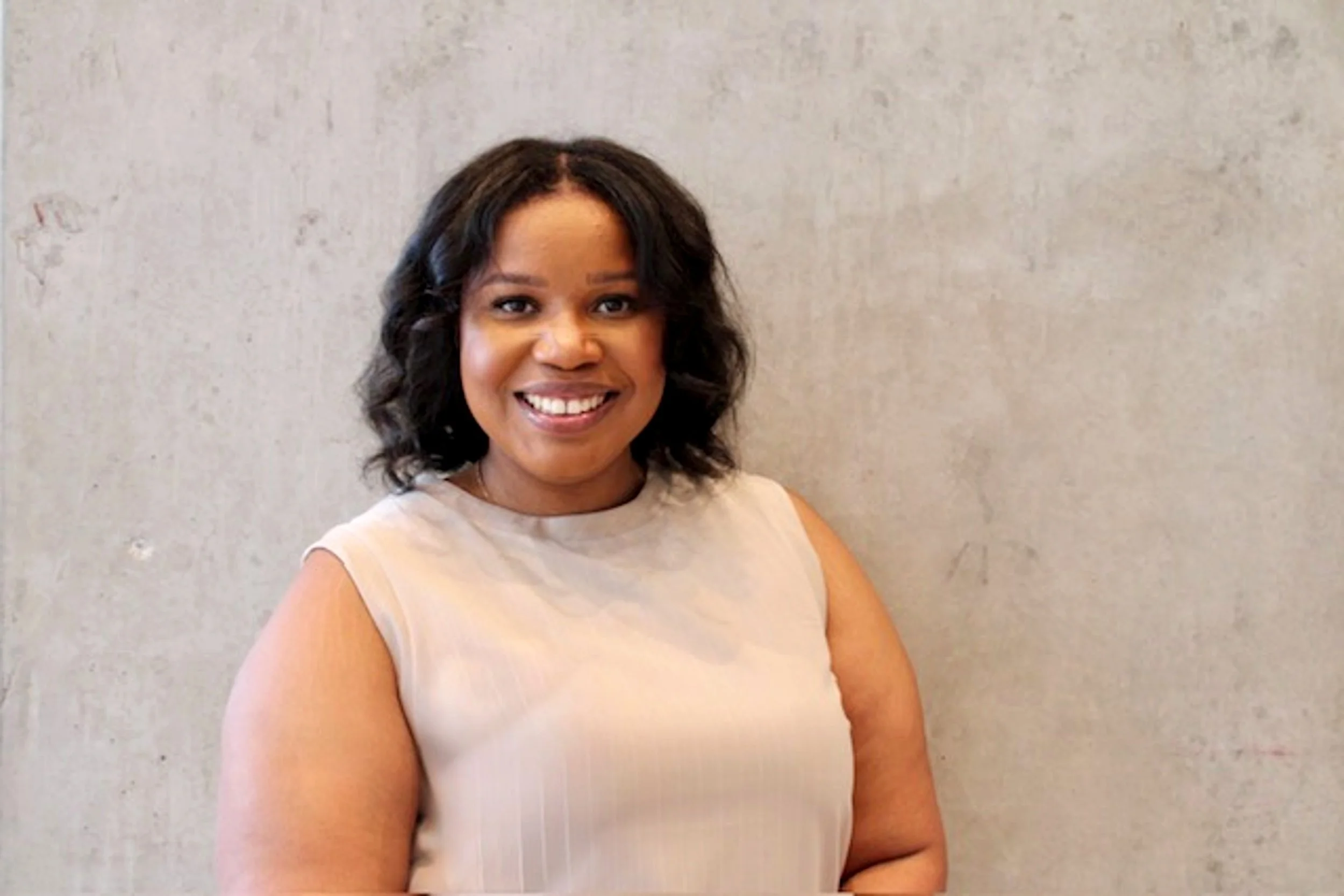A smiling woman with shoulder-length black hair stands against a textured beige wall, wearing a light beige sleeveless top.