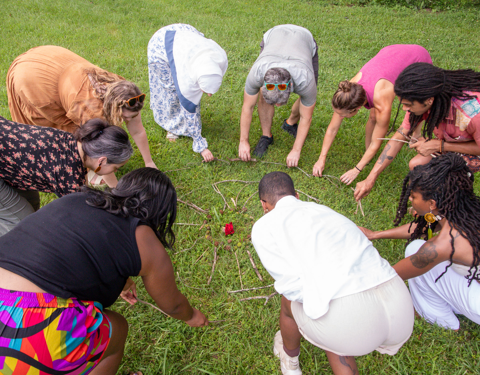 A diverse group of people kneeling on grass in a circle, holding sticks, surrounding a small red flower, participating in a collective activity outdoors.