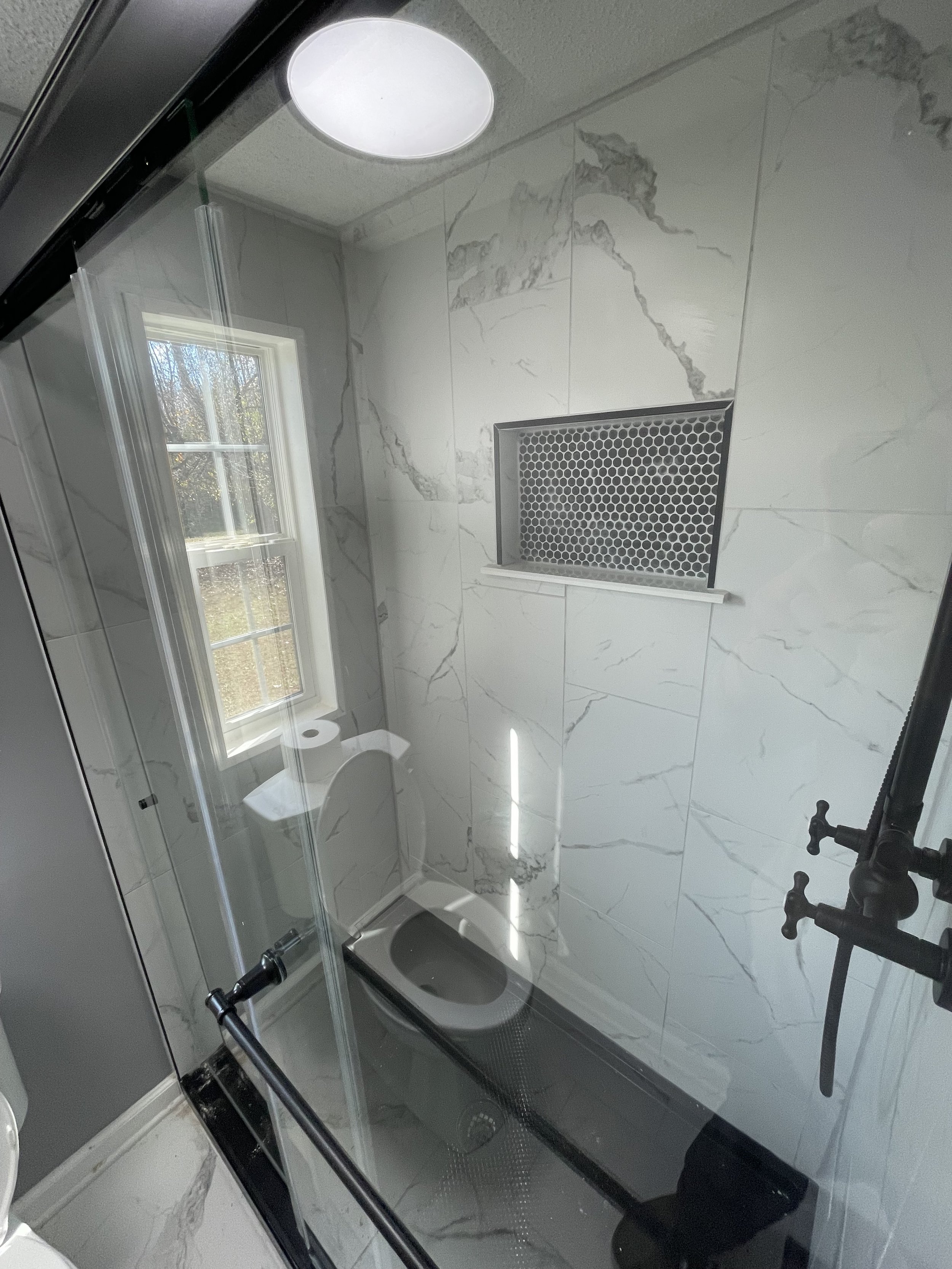 Small bathroom with marble walls, a window, a toilet, and a shower area with black fixtures and a built-in shelf with a black honeycomb-style vent cover.