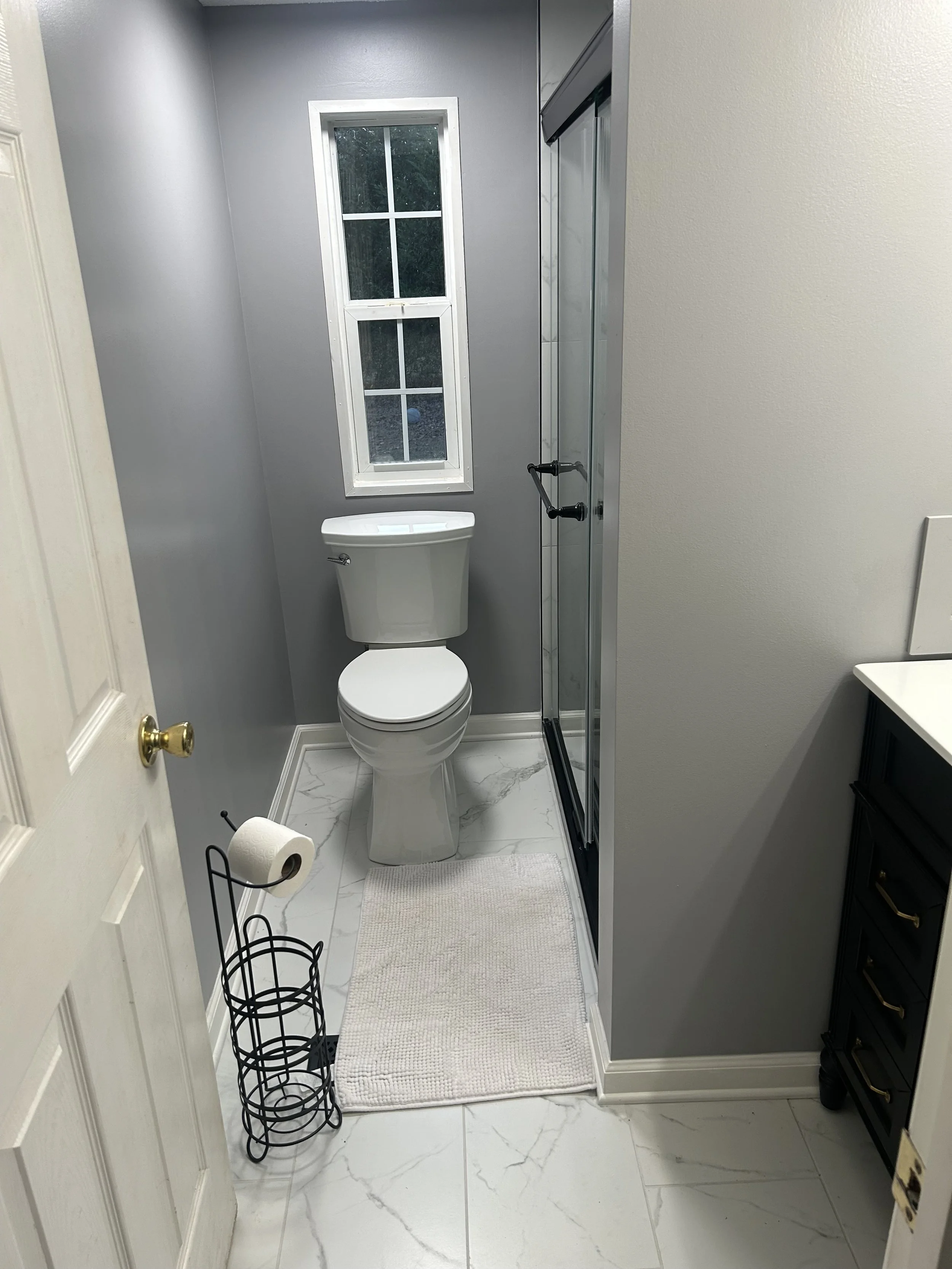 A small bathroom with gray walls, white marble floor, a white toilet, a black metal toilet paper holder, a white bath mat, a small window, a glass shower door, and a black vanity with gold handles.