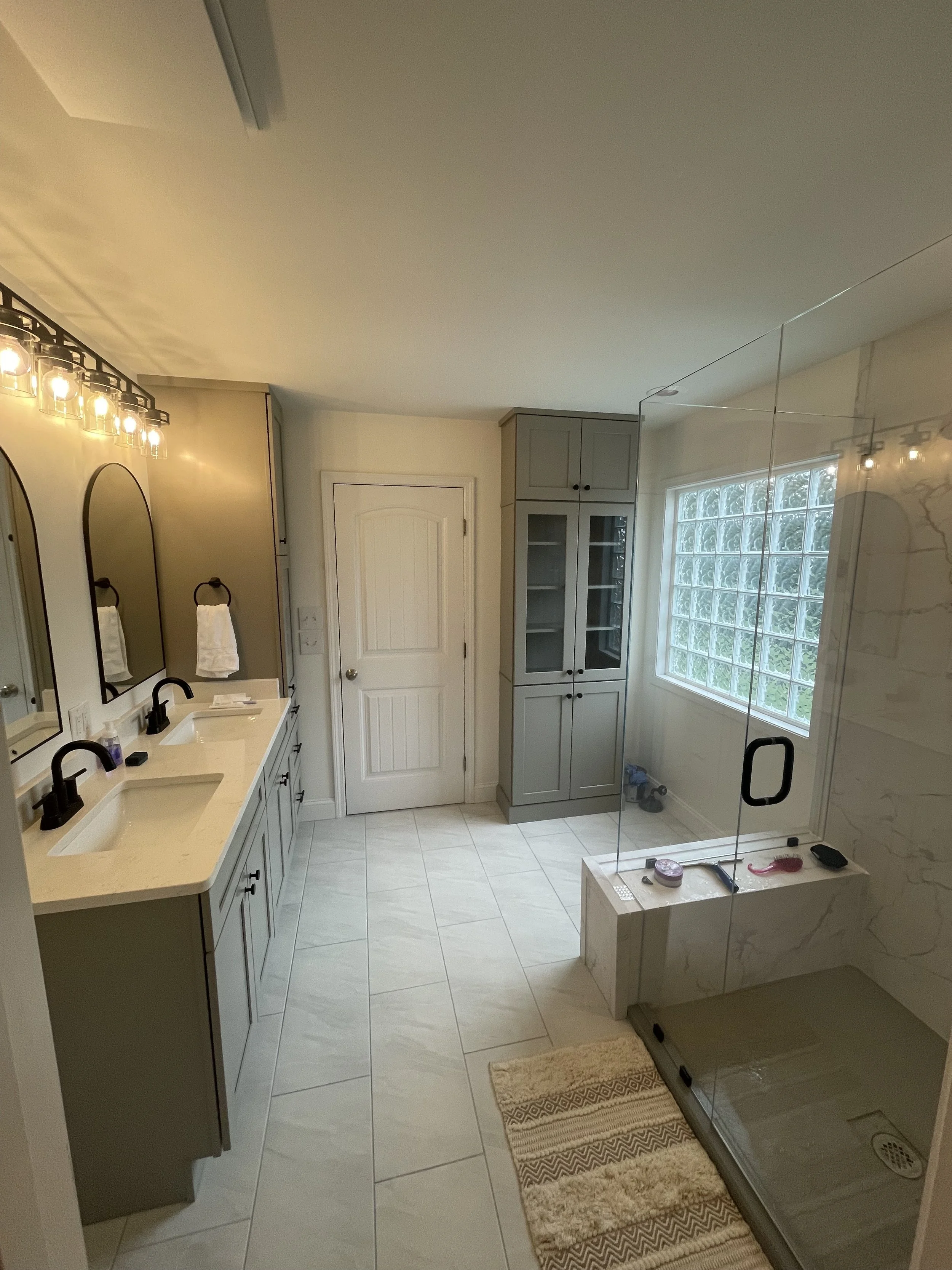 A bathroom with a dual sink vanity, black faucets, large mirror, wall-mounted light fixture, beige towels, a closed closet door, glass shower enclosure, window, and a small rug in front of the shower.