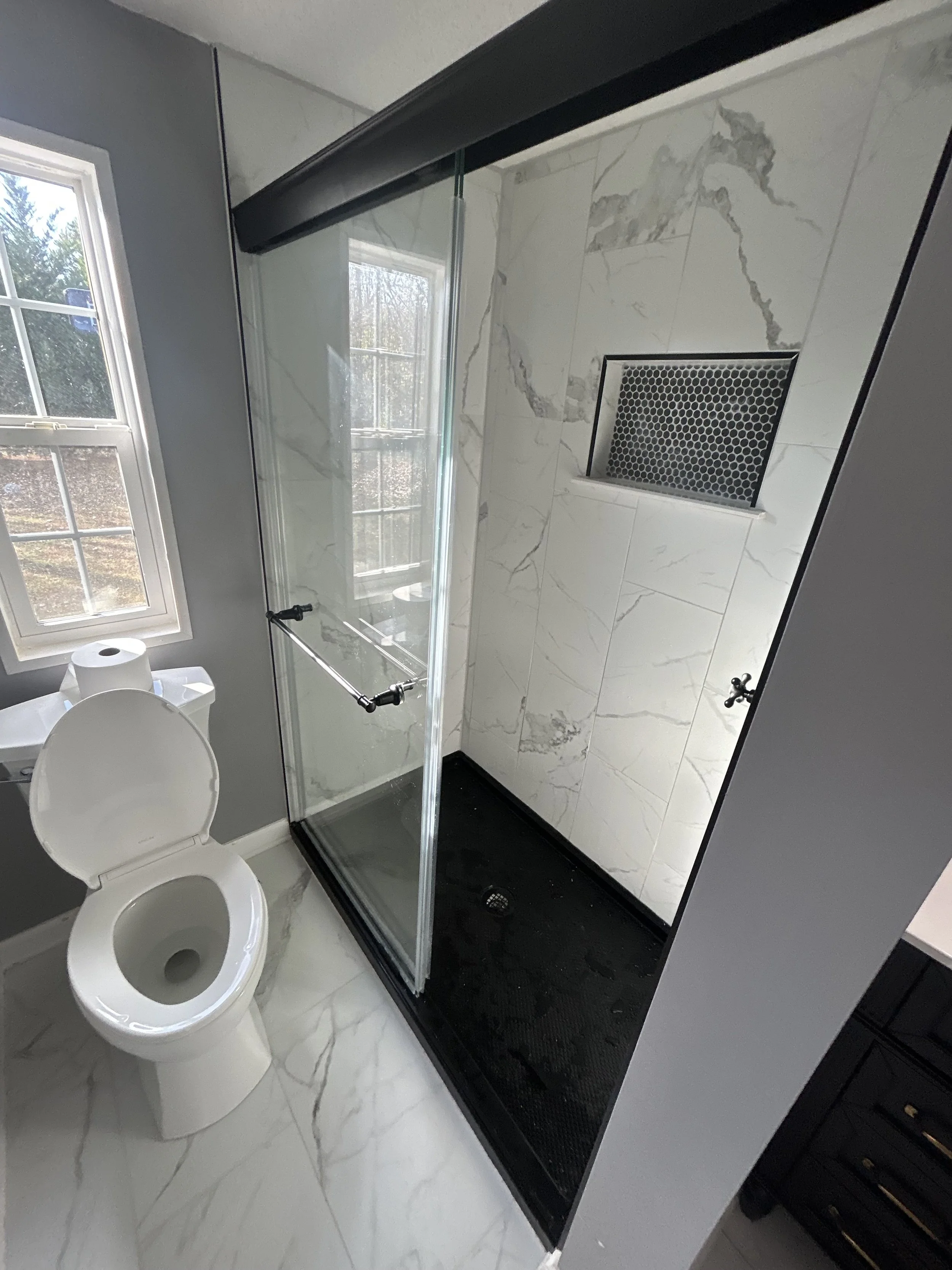 Bathroom with toilet, window, and a walk-in shower with marble tiles and a black shower drain.