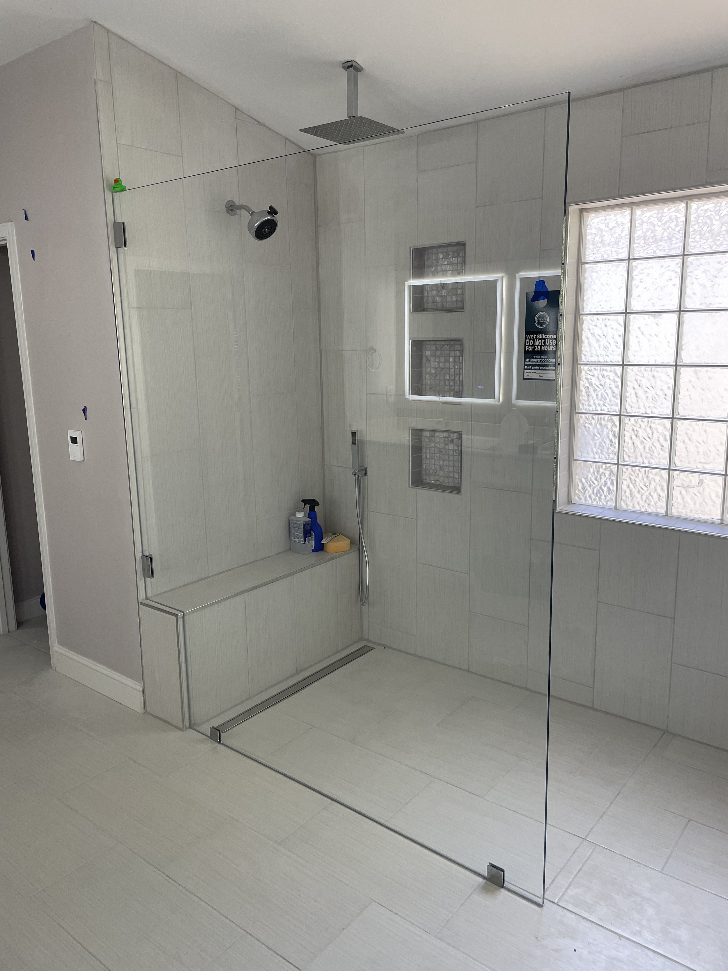 Modern walk-in shower with glass partition, white tiled walls, a rain showerhead, a handheld shower, built-in shelves, and a window with textured glass.