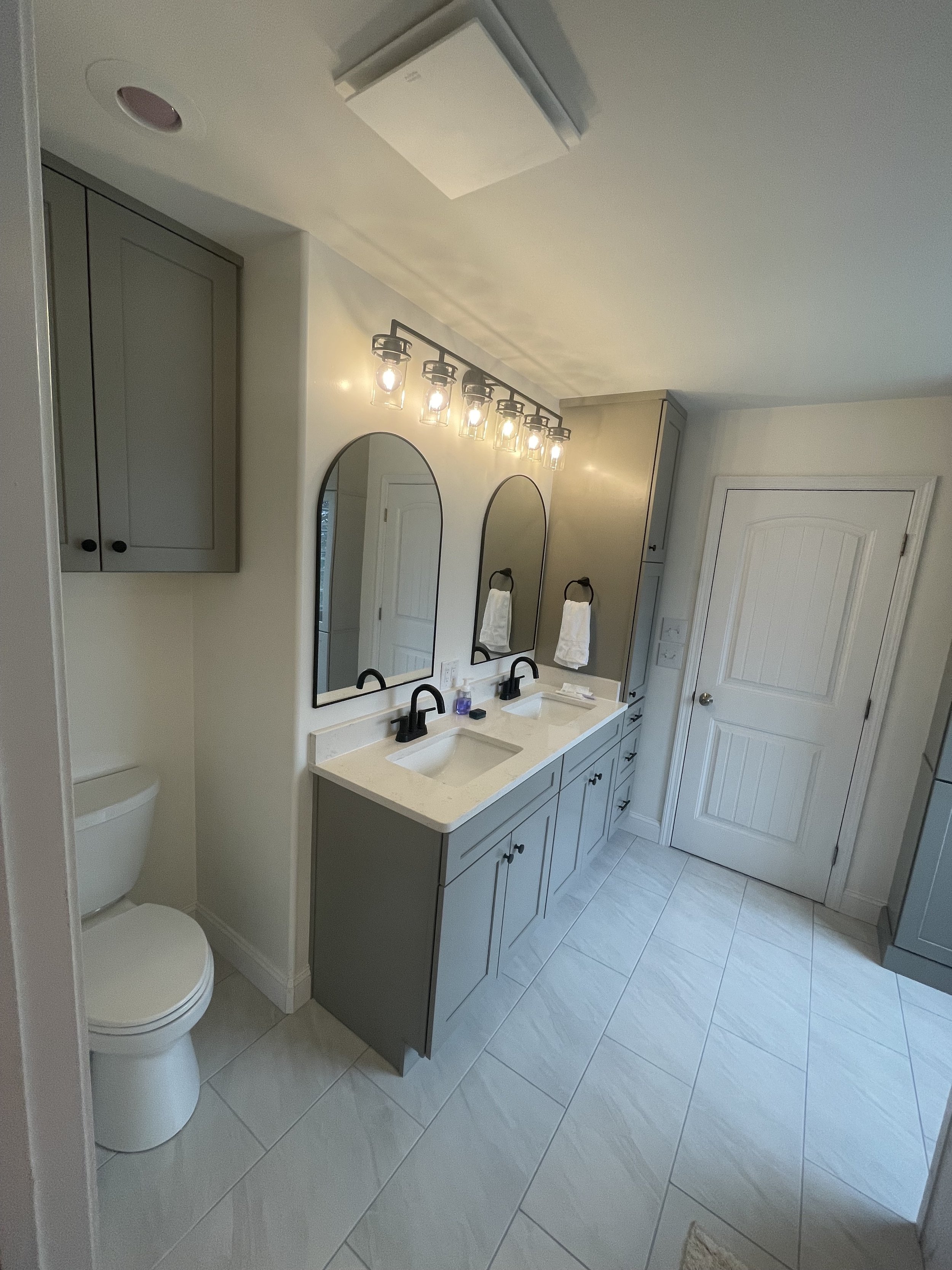 Bathroom with a double vanity sink, two mirrors, gray cabinets, black faucets, towel racks with white towels, a ceiling light fixture, and a toilet beside the vanity.
