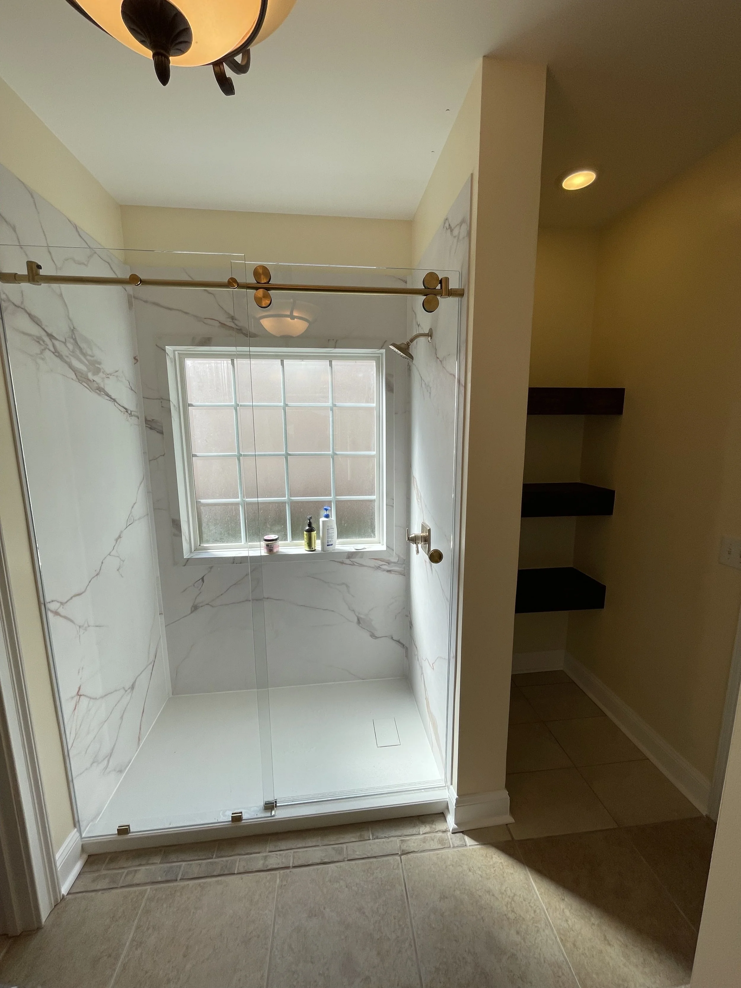 A bathroom with a glass shower enclosure, marble walls, a window, bottles of soap and shampoo on the windowsill, and beige tiled flooring.