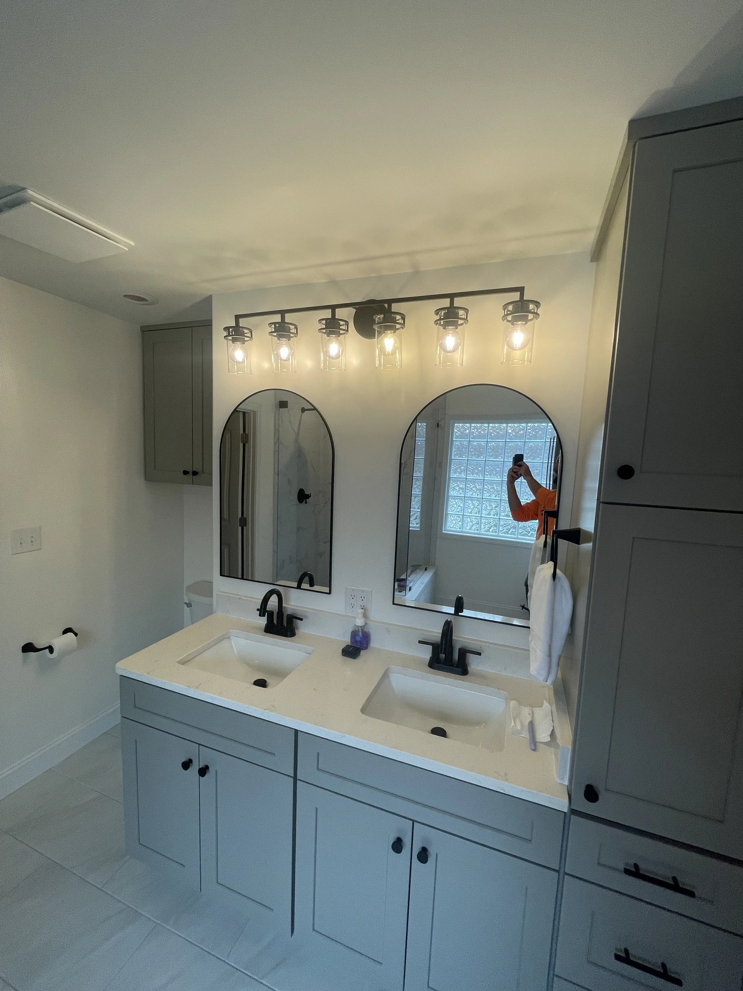 A bathroom vanity with double sinks, gray cabinets, black fixtures, and a large mirror above. There are wall cabinets and a person taking a photo reflected in the mirror. There are two black handles on towels and a window in the background.
