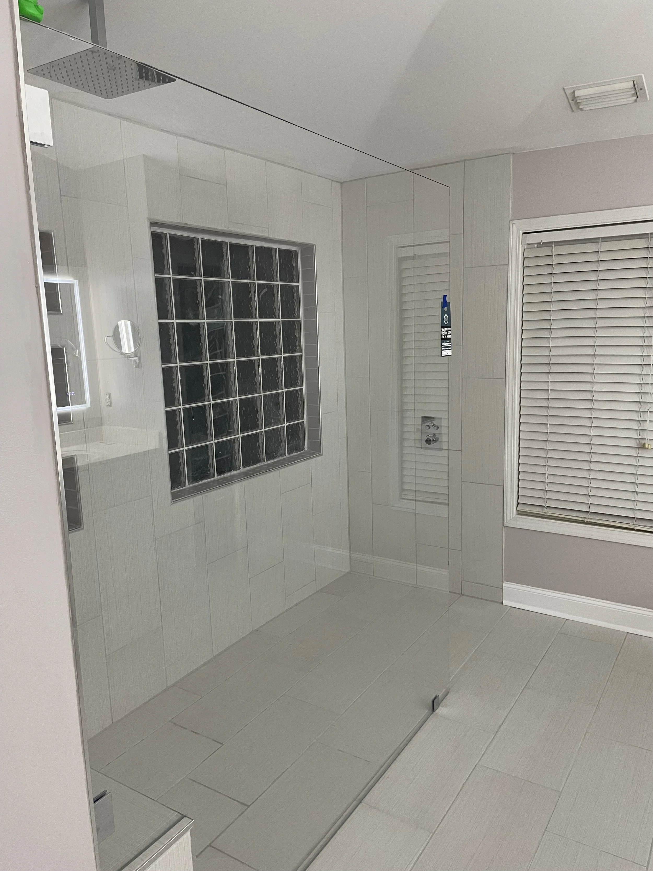 A modern bathroom with a glass shower enclosure, a square showerhead on the ceiling, a frosted glass window made of glass blocks, and a window with closed white blinds. The walls and floor are tiled in light colors.