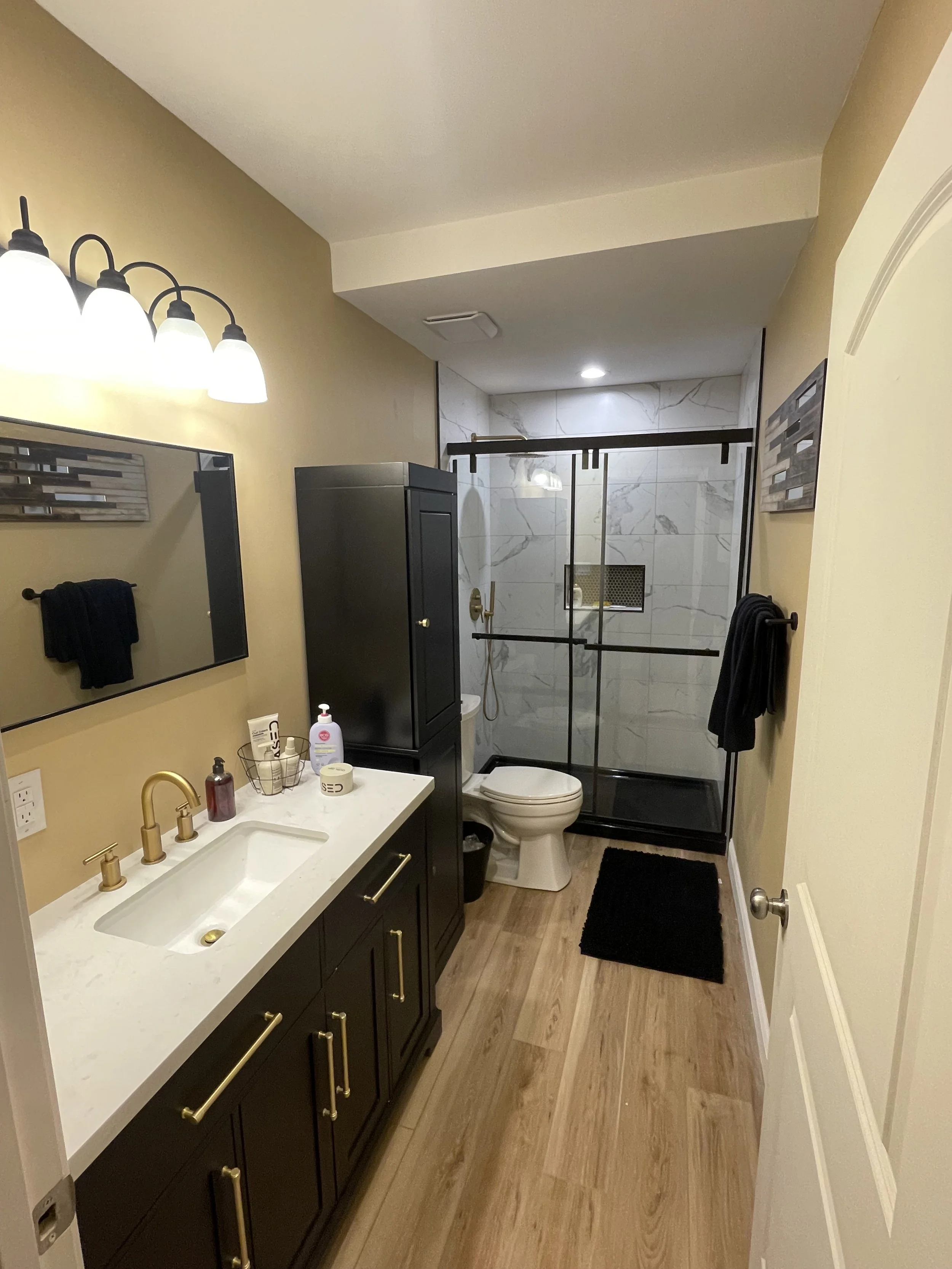 A modern bathroom featuring a white vanity with gold handles, a black cabinet, a toilet, a glass-enclosed walk-in shower with marble tiles, a wall mirror, and black towels hanging on the wall.