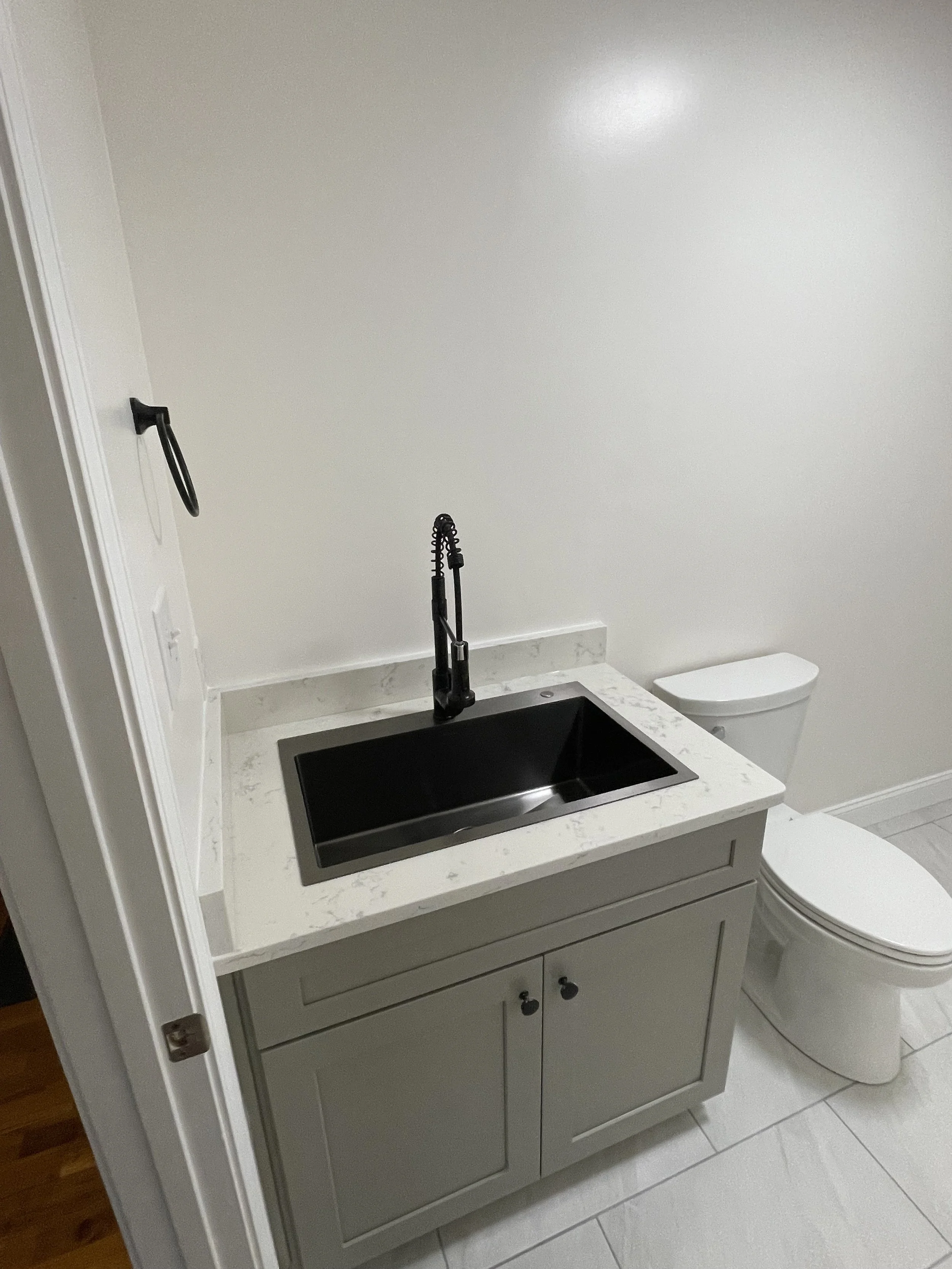 A bathroom with a gray vanity with a white marble countertop and black sink, a black gooseneck faucet, a toilet, and a plain white wall.