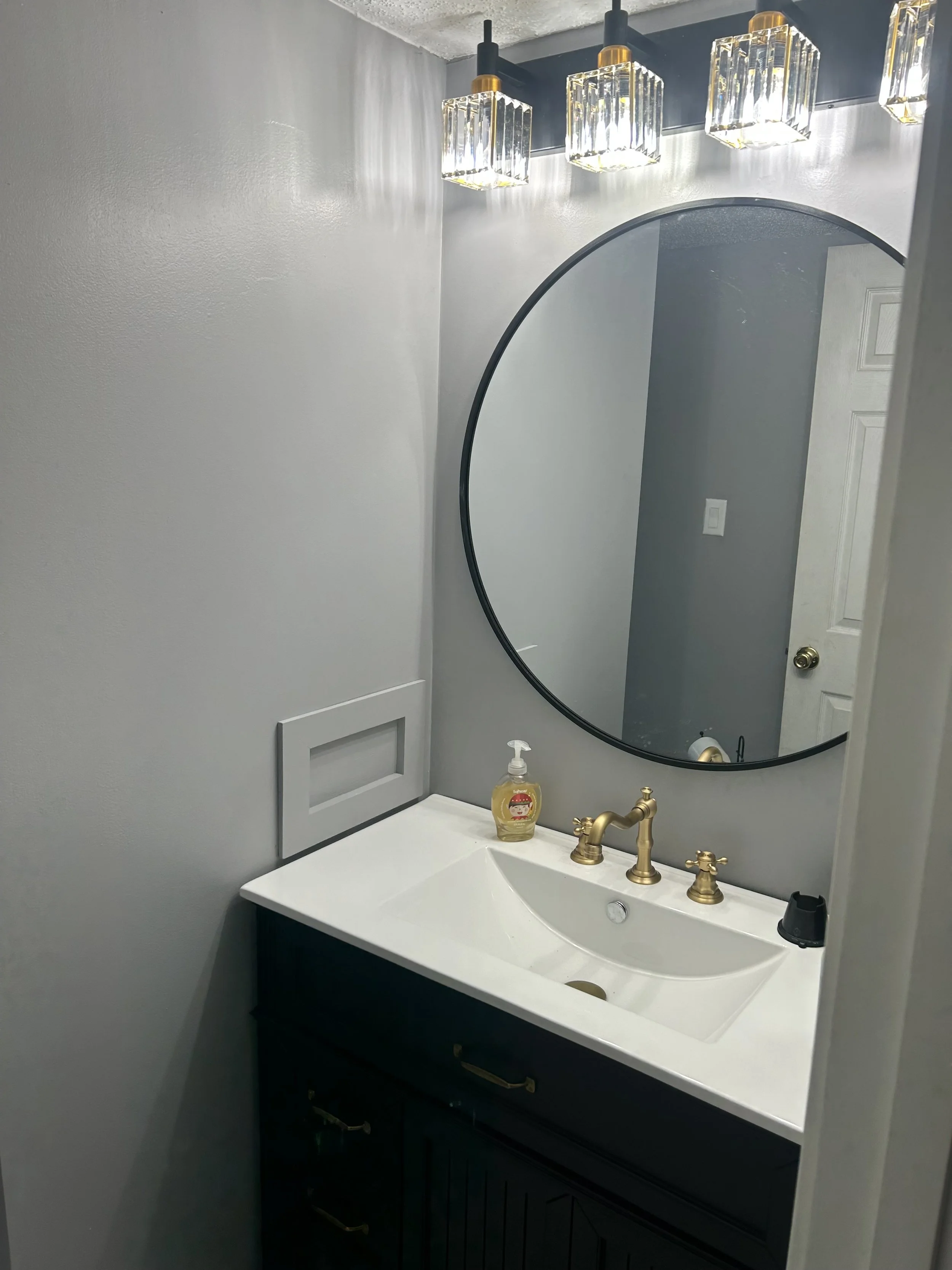 A bathroom vanity with a white sink, gold fixtures, a black cabinet, a round mirror, and a row of four modern light fixtures with glass shades above the mirror.