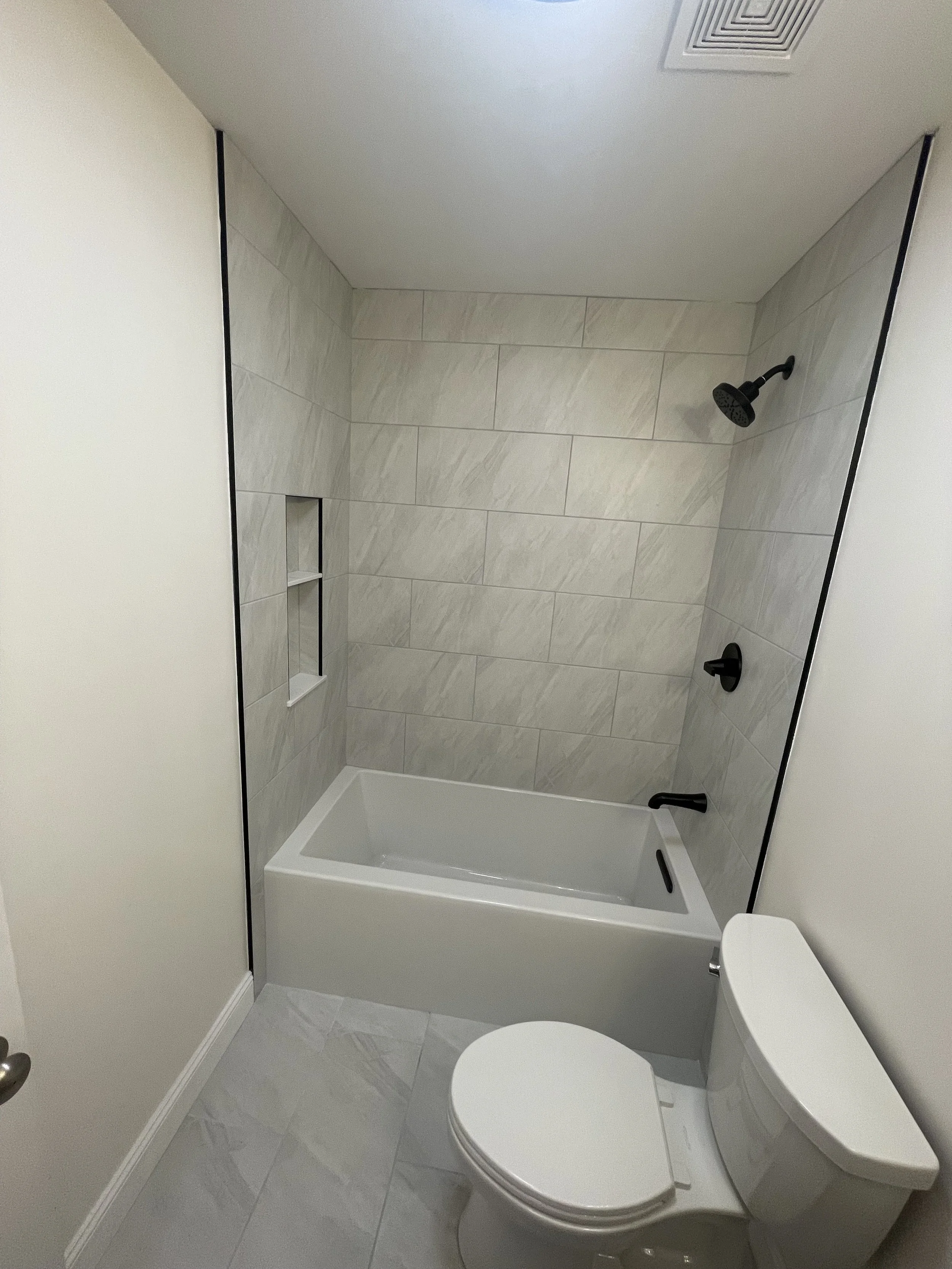A bathroom with a white toilet, glass shower door, tiled shower area with wall niche, black fixtures, and a white ceiling with a vent.